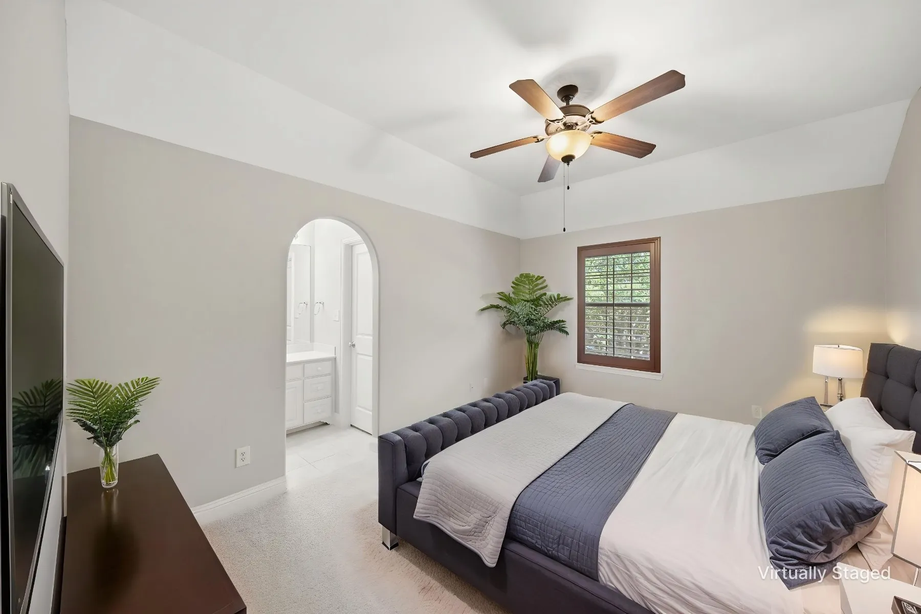 Tray ceiling bedroom featuring a ceiling fan with integrated lighting, a window with wood-frame trim, and an arched doorway leading to an additional space with white cabinetry