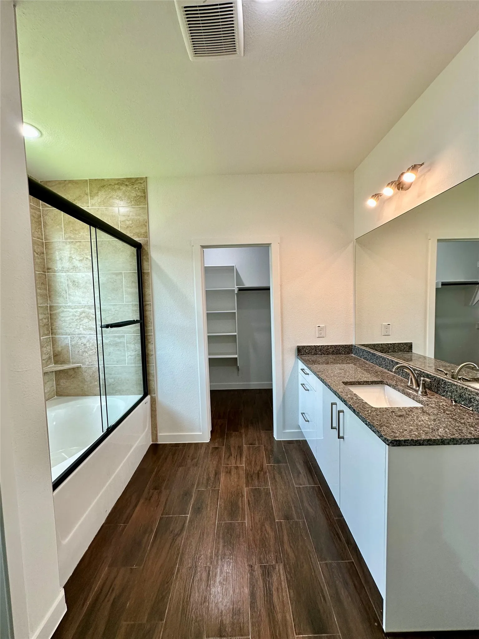 Bathroom featuring a wood-finish tile floor, a shower-tub combination with a tiled surround and glass enclosure, and a double vanity with stone countertops and undermount sinks