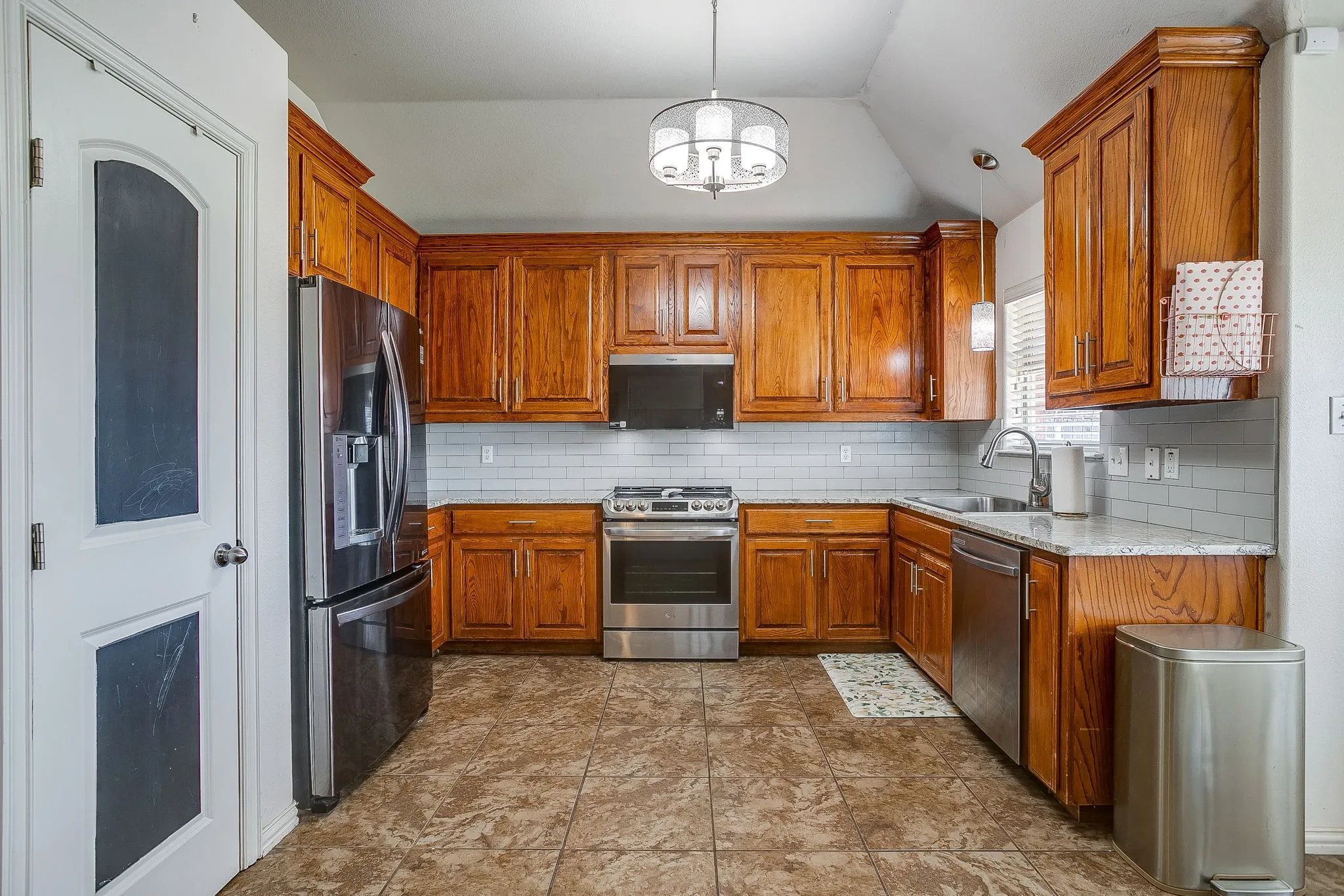 Kitchen featuring wood-finish cabinetry, stainless steel appliances, a white subway tile backsplash, light-toned countertops, and patterned floor tiling