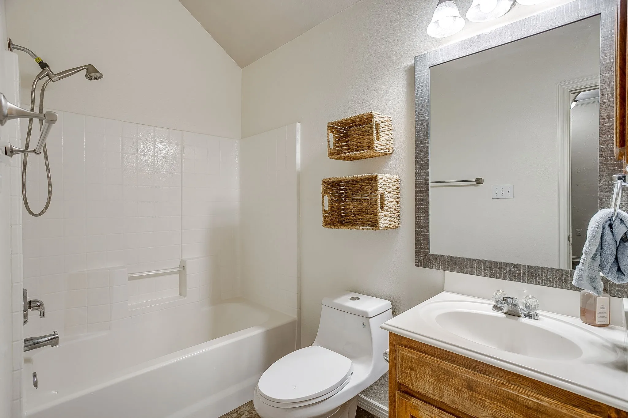 Bathroom featuring a white tub and shower combination with a grab bar, a single vanity with a white countertop and wood-finish cabinetry, a framed mirror, and vaulted ceilings