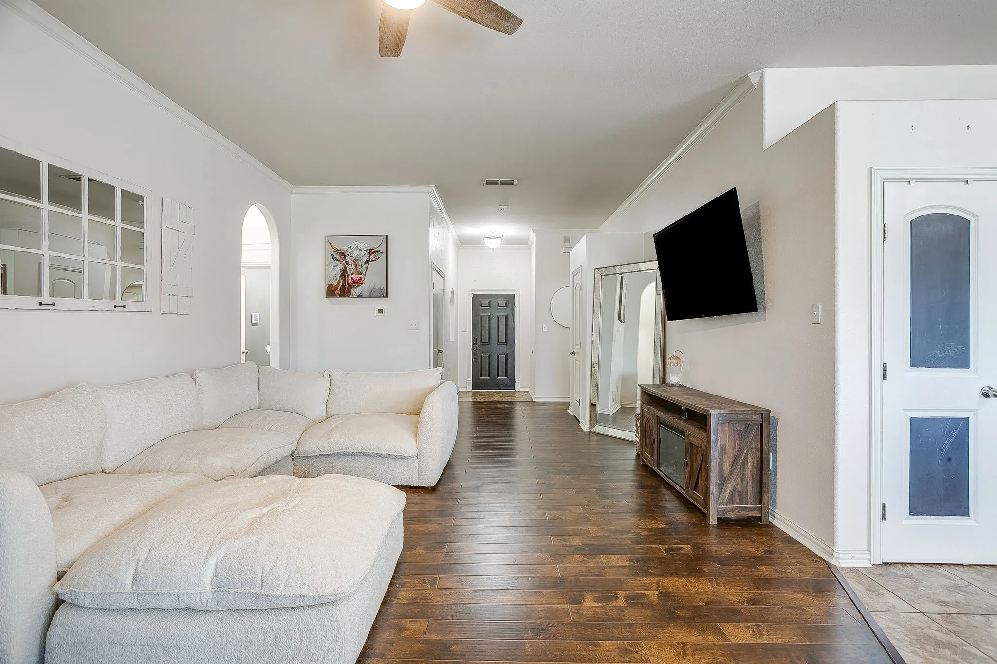 Spacious living area featuring rich wood-finish flooring, crisp white walls, a ceiling fan, and an arched entryway
