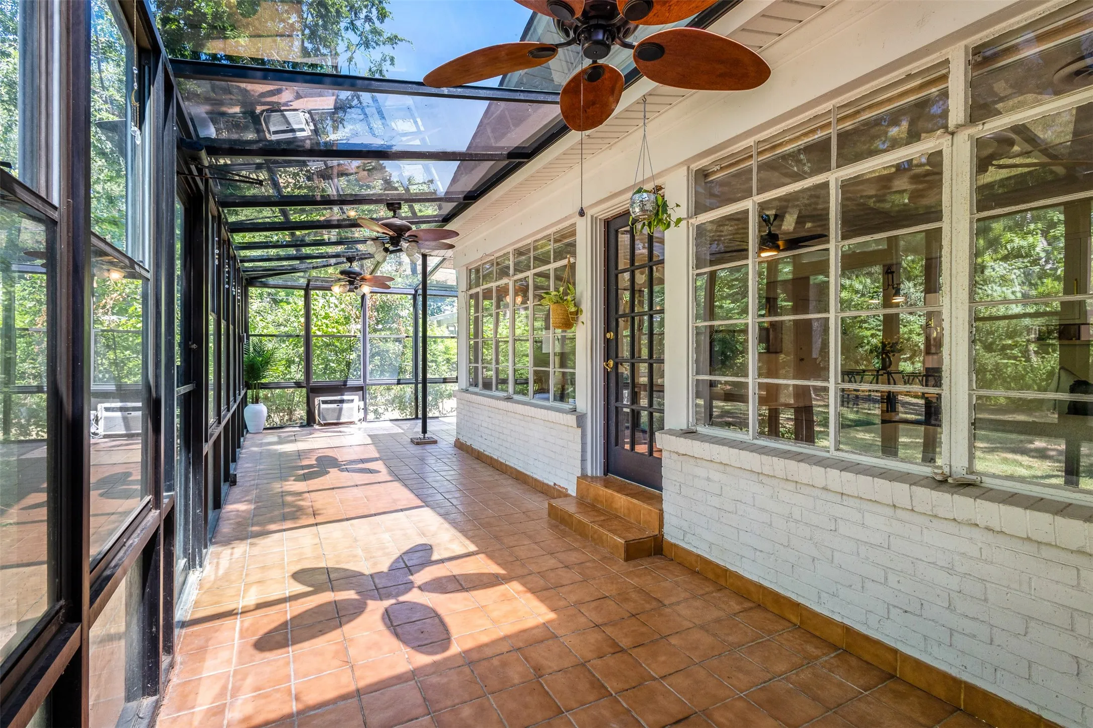Expansive sunroom featuring a glass roof, full-height glass walls, terracotta tile flooring, multiple ceiling fans, and a white painted brick exterior wall