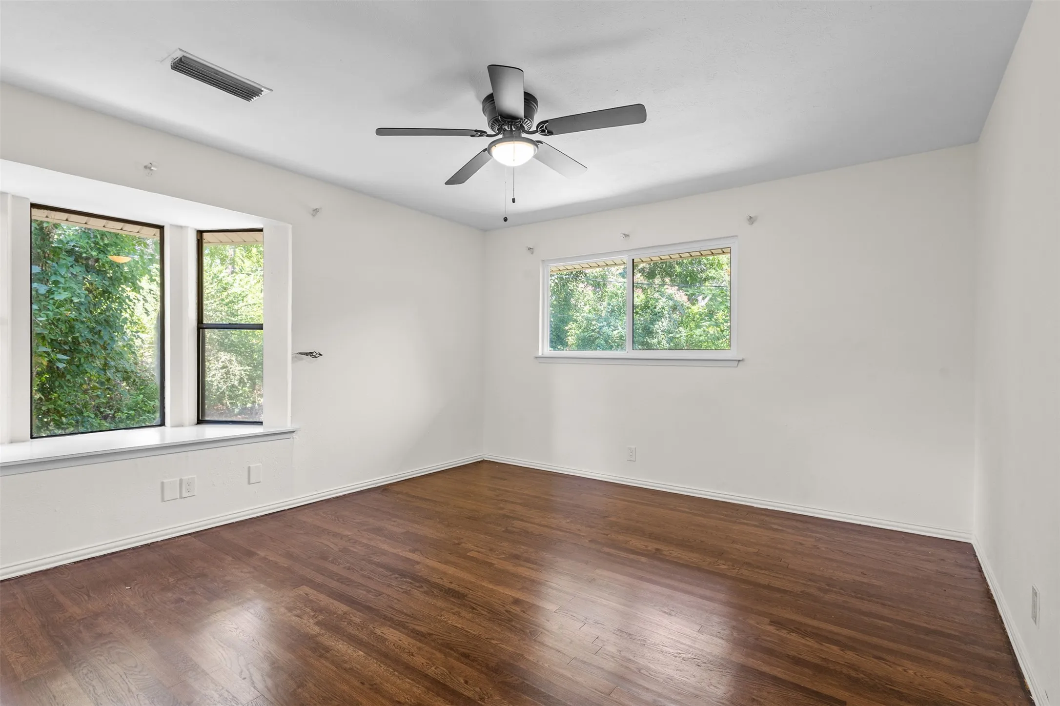 Spacious room featuring wood-finish flooring, a bay window with a sill, a ceiling fan with integrated lighting, and multiple windows