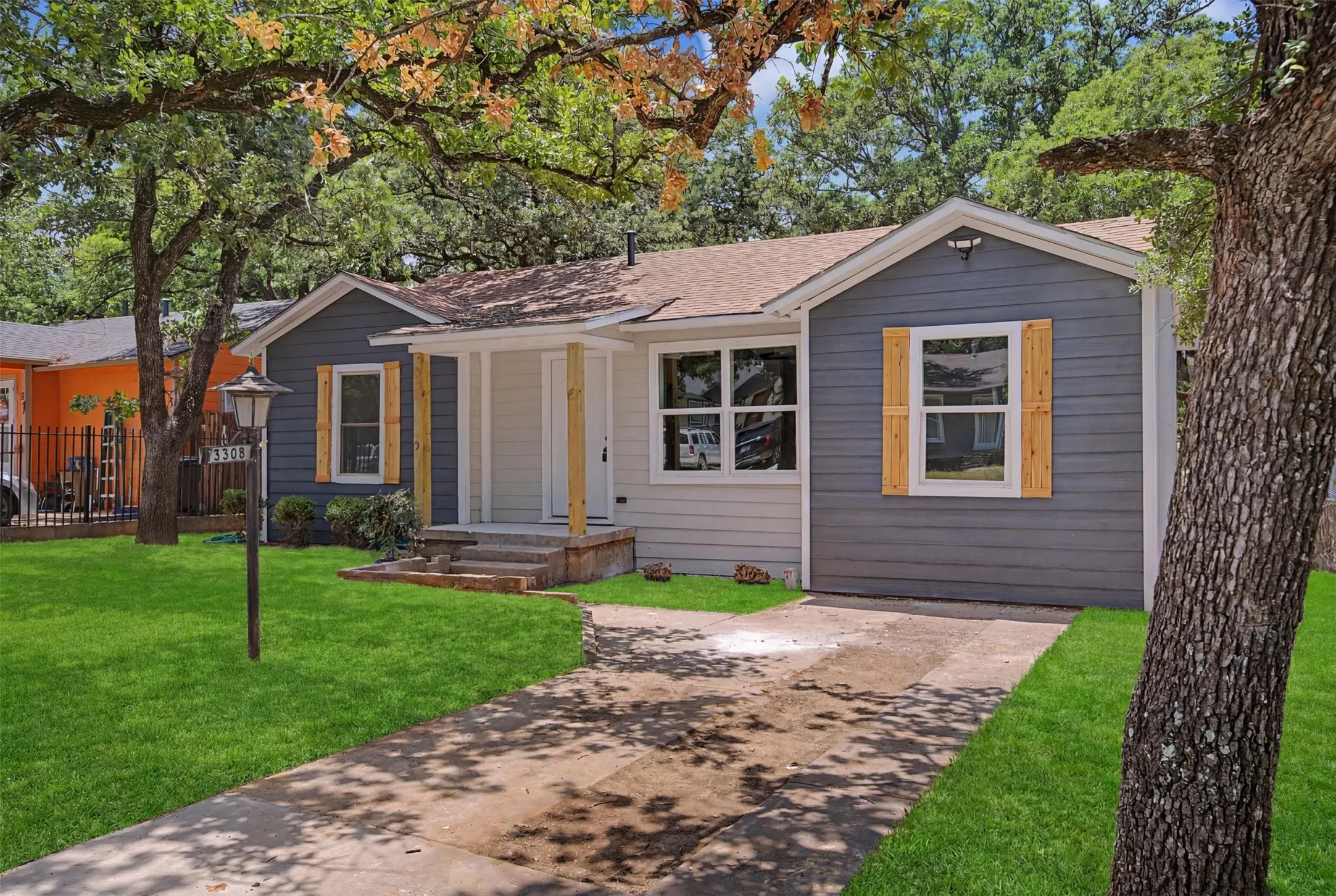 Single-story residence featuring a gray and light-colored siding exterior, natural wood window shutters, a covered front porch with natural wood pillars, a concrete driveway, and a front yard with mature trees