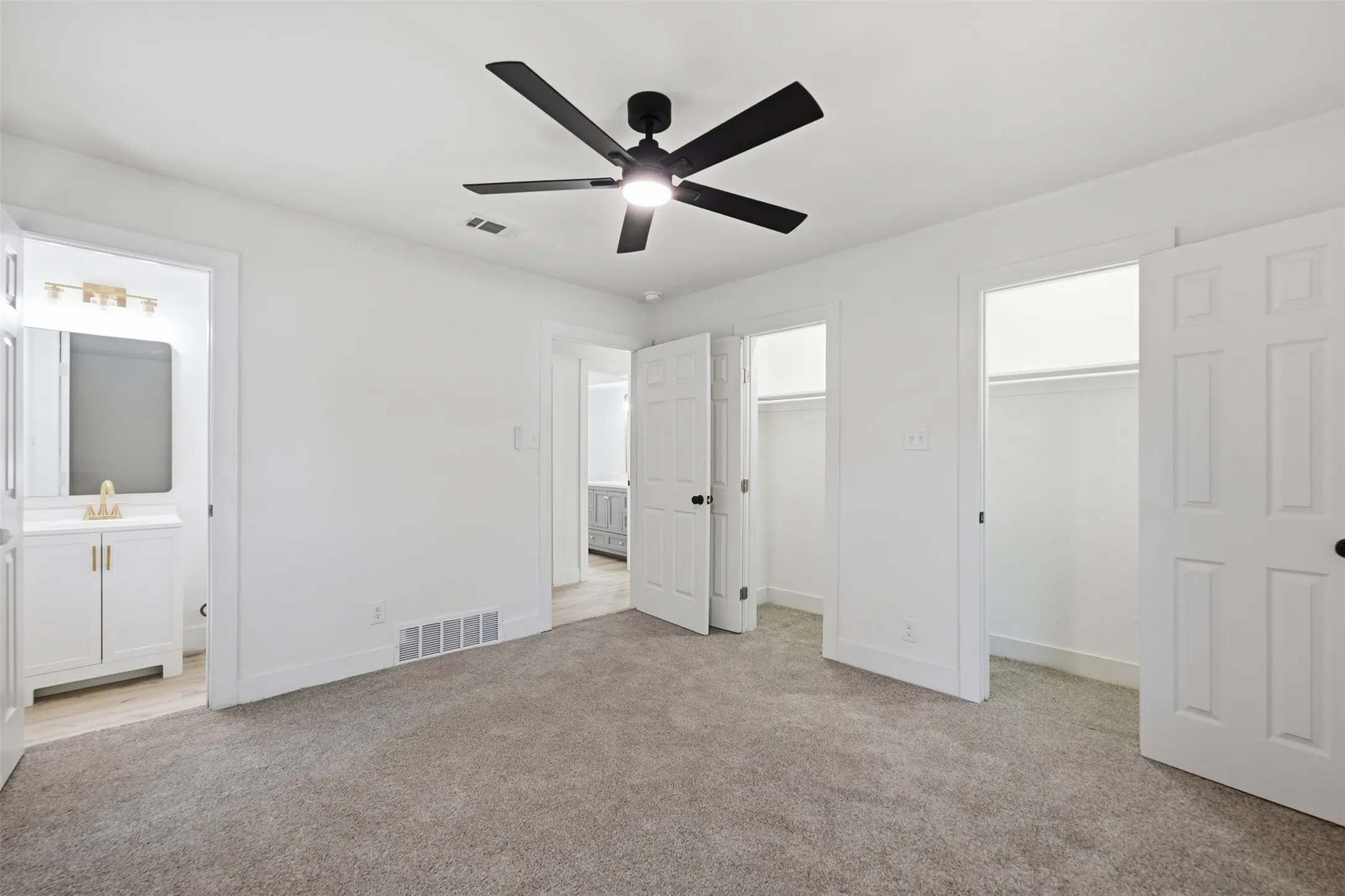 Carpeted room featuring a matte black ceiling fan, two closet openings with hanging rods, and a white interior door leading to a bathroom with a vanity, mirror, and light fixture