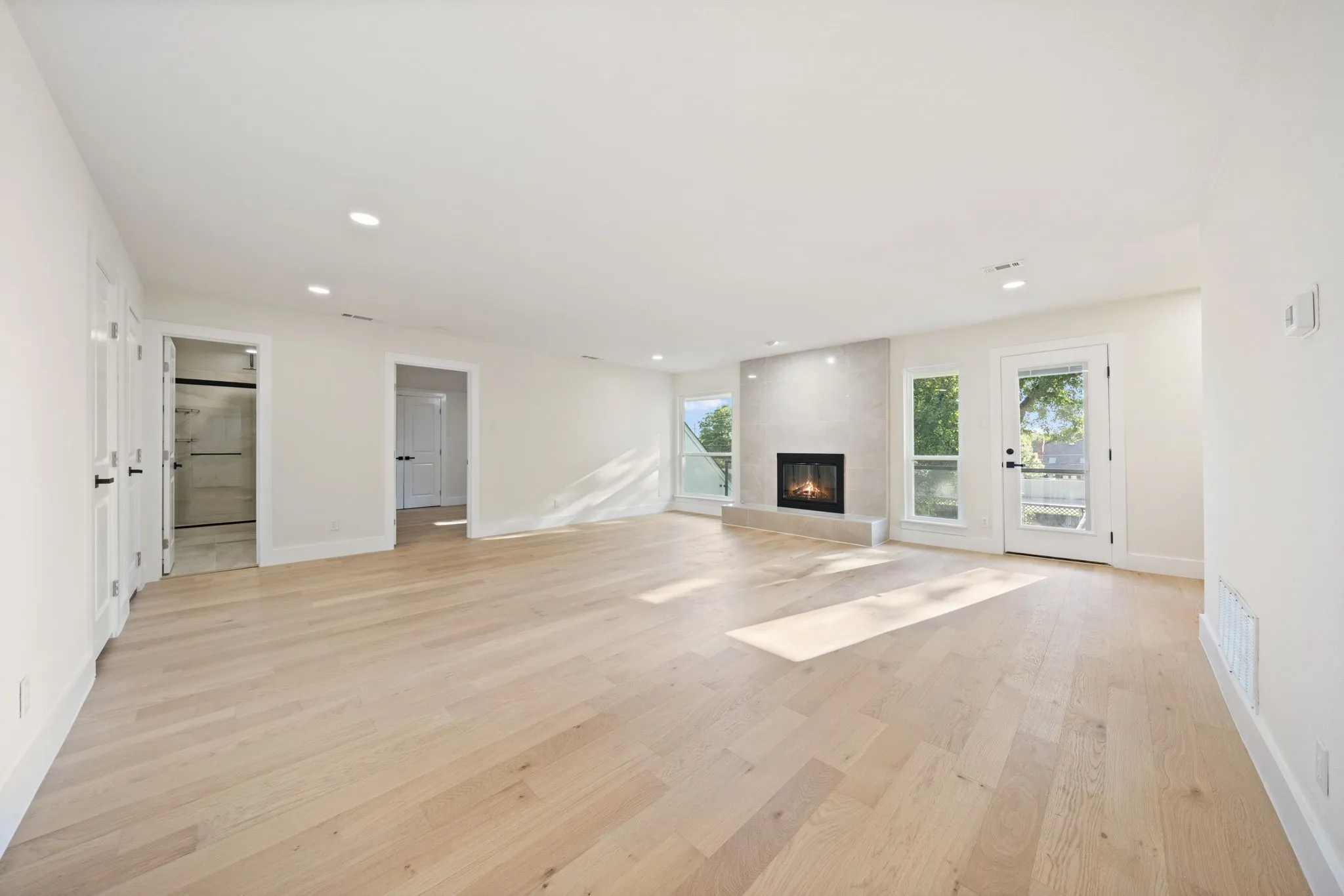 Spacious room with light wood-finish flooring, recessed lighting, a built-in fireplace with a stone surround, and multiple windows and an exterior door providing natural illumination