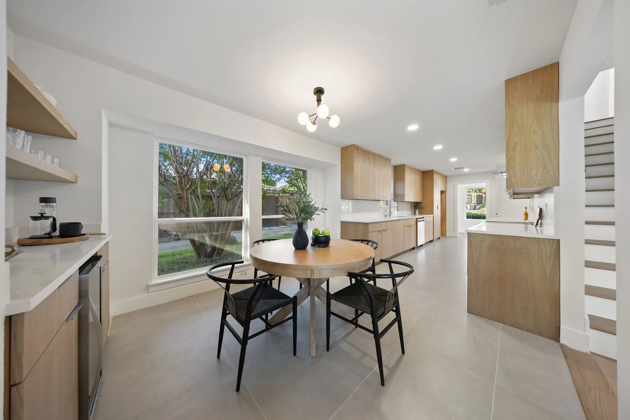 Open-concept kitchen and dining area featuring light wood-finish cabinetry, light gray tile flooring, recessed lighting, and a modern chandelier