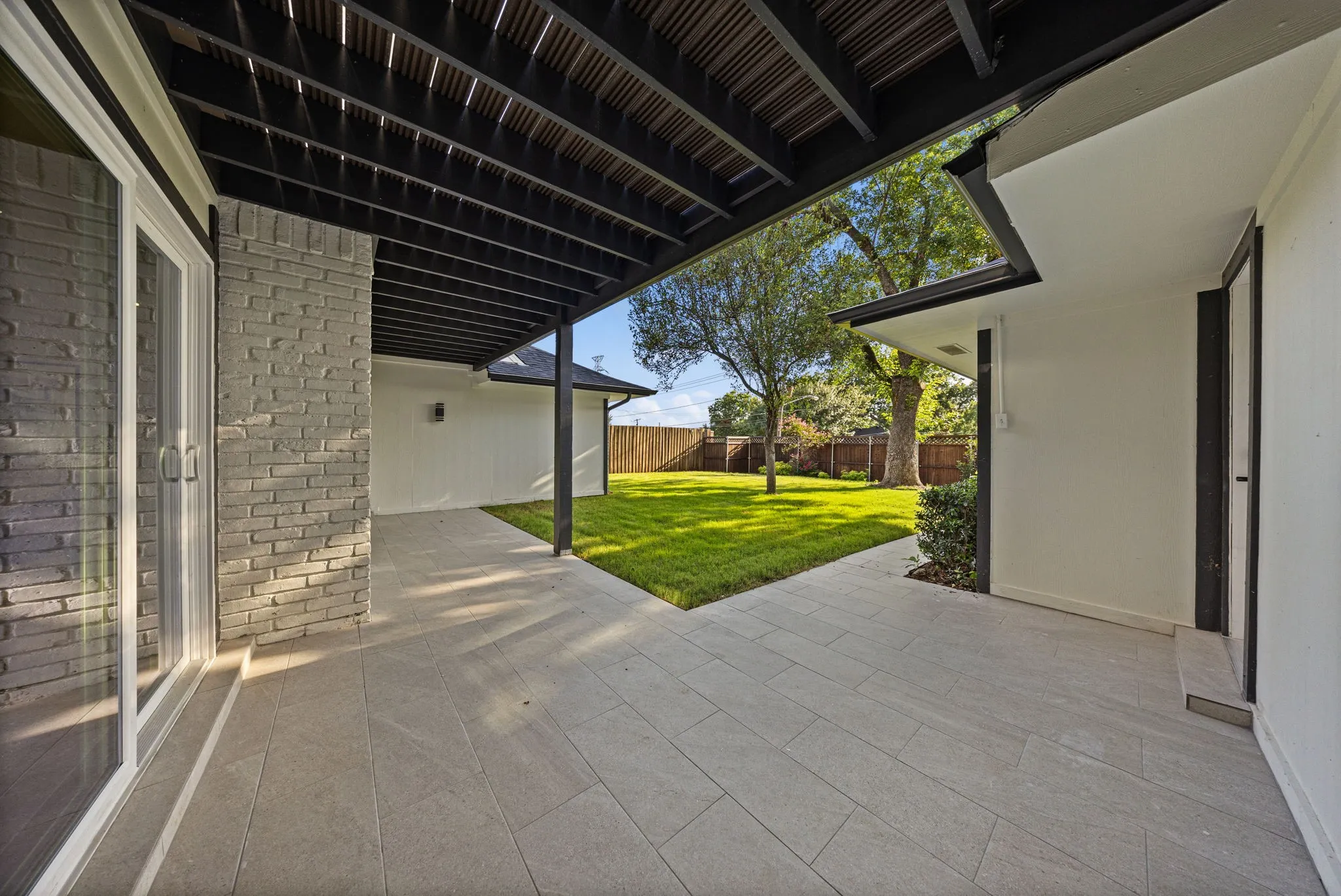 Expansive outdoor living area featuring a covered patio with dark wood ceiling beams, light grey plank-style flooring, a grey brick exterior wall, and a sliding glass door