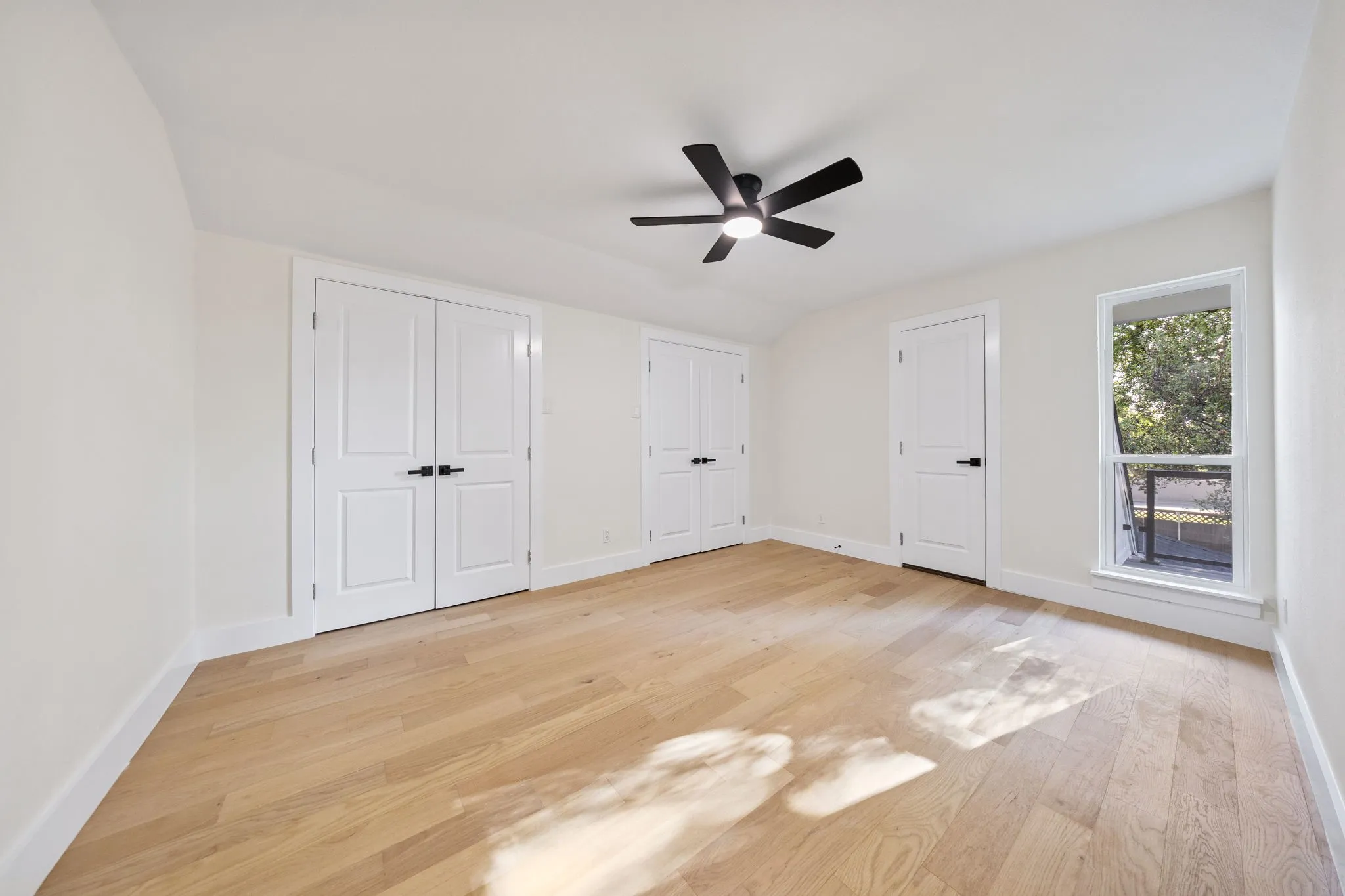 Bright room featuring wood-finish flooring, a contemporary ceiling fan, white paneled doors with dark hardware, and a large window with white trim