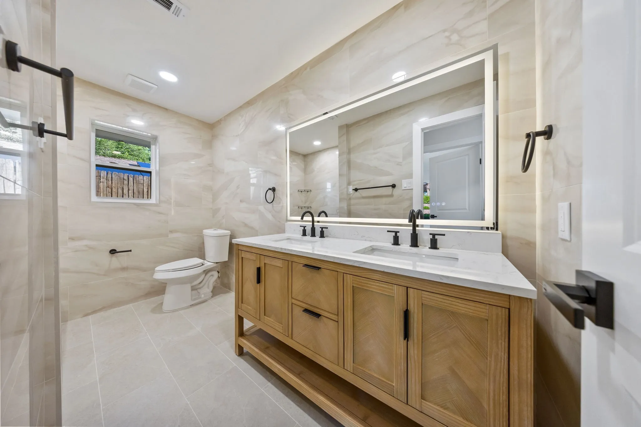 Bathroom featuring a light wood-finish double vanity with white countertop, matte black fixtures, and an illuminated mirror