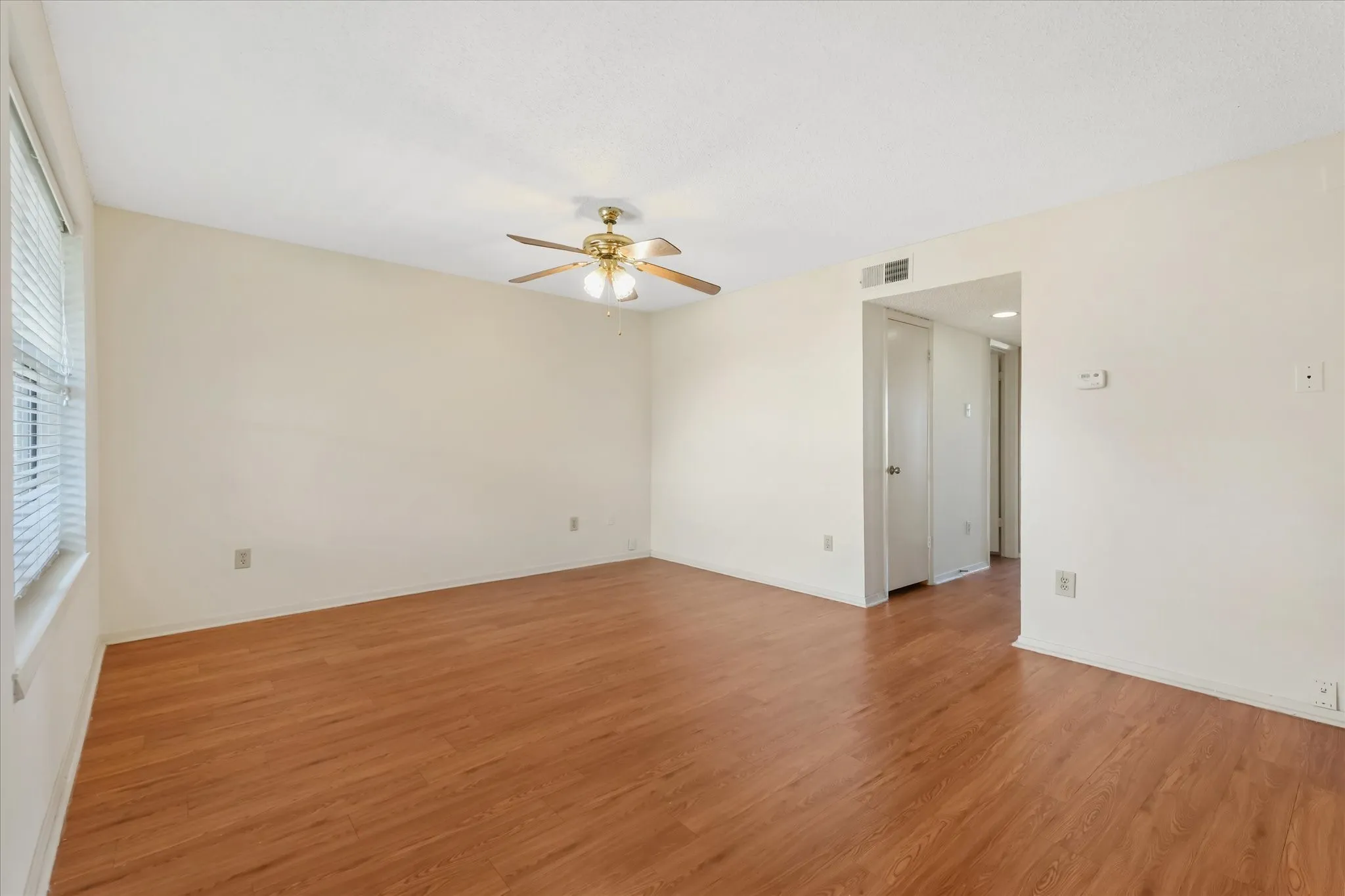 Spacious interior room featuring wood-finish flooring, light-colored walls, a ceiling fan with integrated lighting, and a window with horizontal blinds