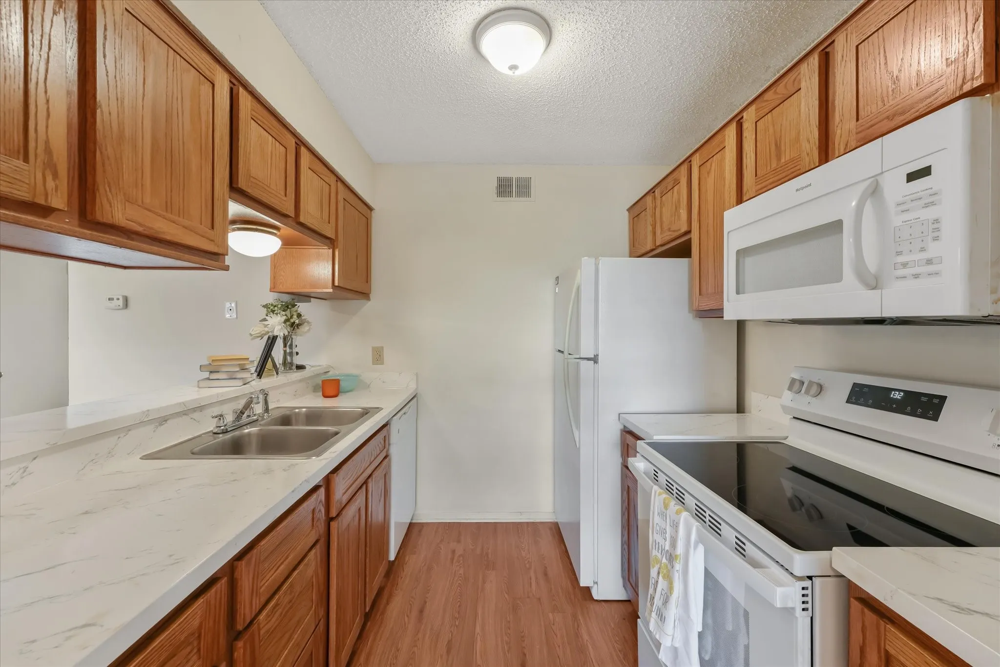 Galley kitchen featuring wood cabinetry, white countertops, stainless steel double basin sink, and white appliances including a refrigerator, range, and built-in microwave