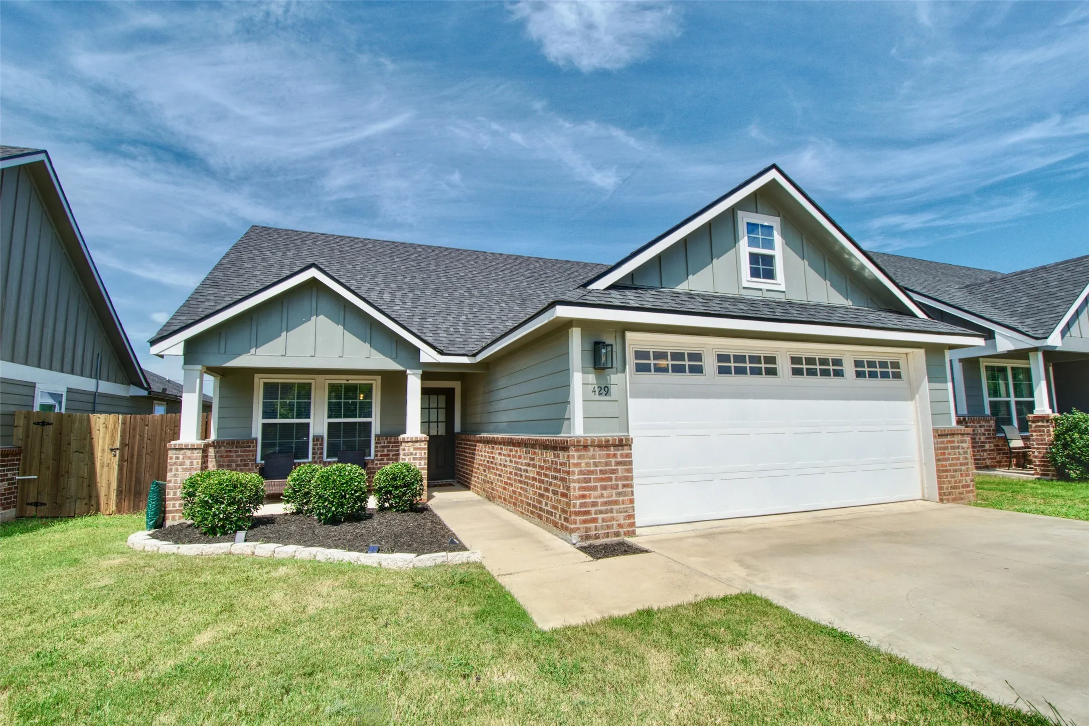 Craftsman-style exterior featuring a covered front porch, brick wainscoting, a two-car garage with transom windows, and a landscaped front yard