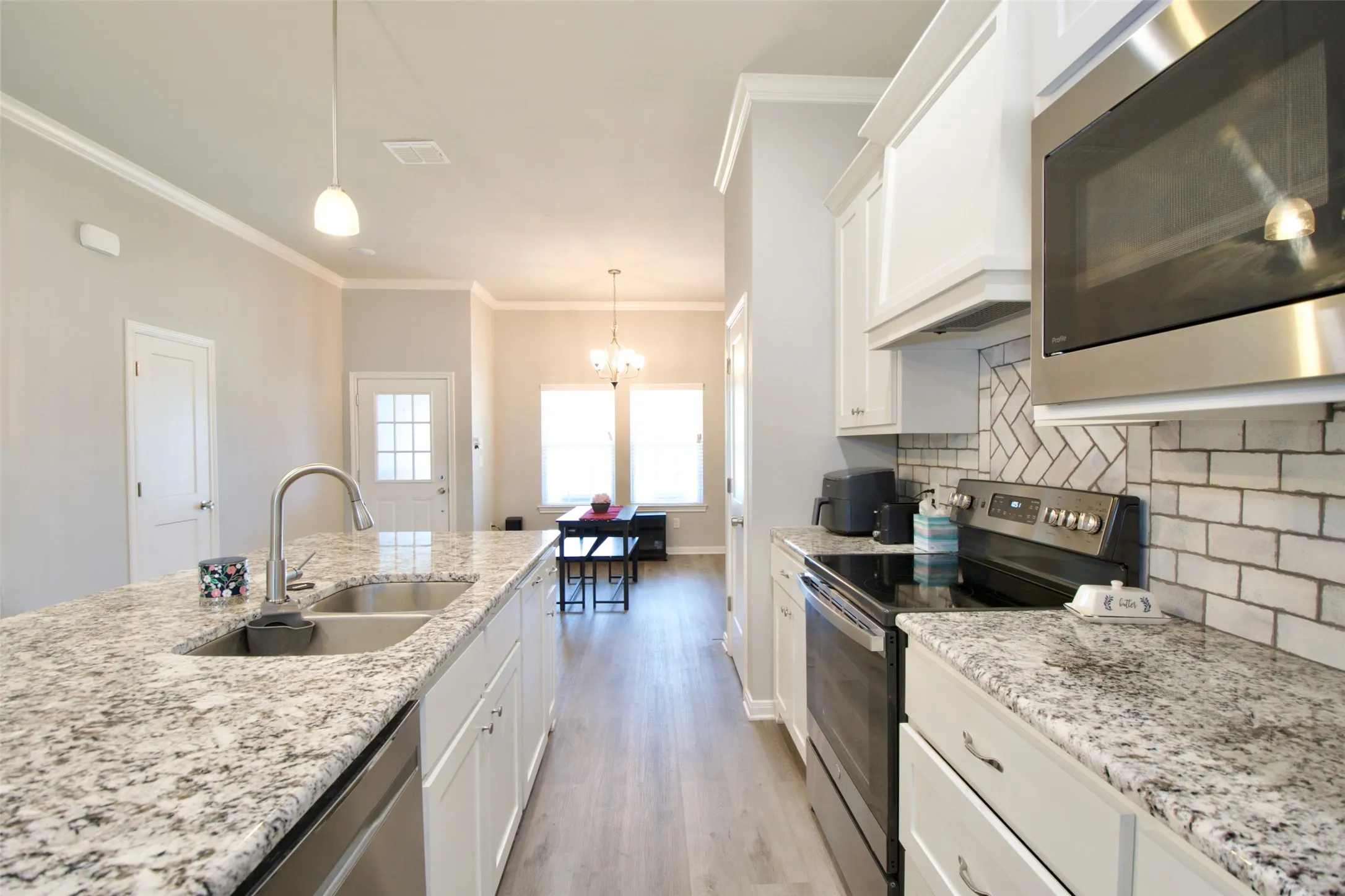 Kitchen featuring an expansive island with a double basin sink, stainless steel appliances, light granite countertops, white cabinetry, and a herringbone pattern tile backsplash