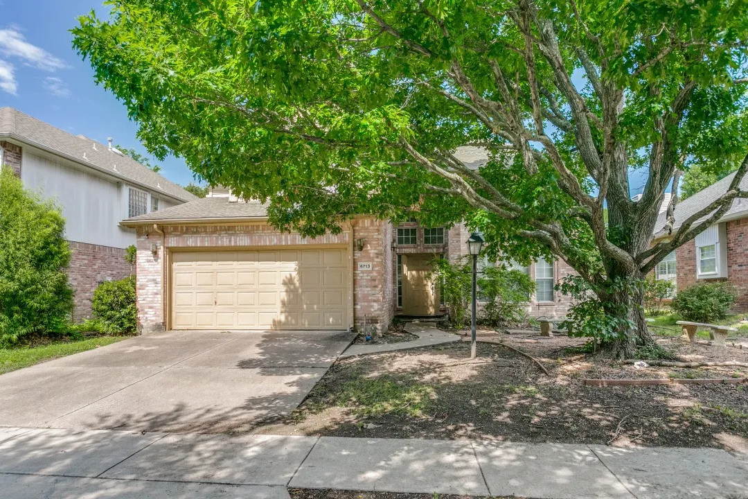 Brick facade residence with an attached garage, featuring a mature tree, concrete driveway, and front sidewalk