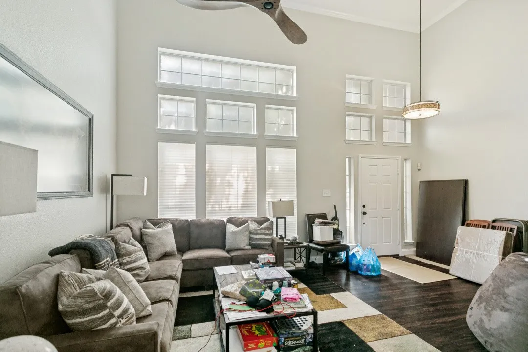 Expansive living space featuring high ceilings, a prominent ceiling fan, dark wood-finish flooring, and multiple large windows with blinds