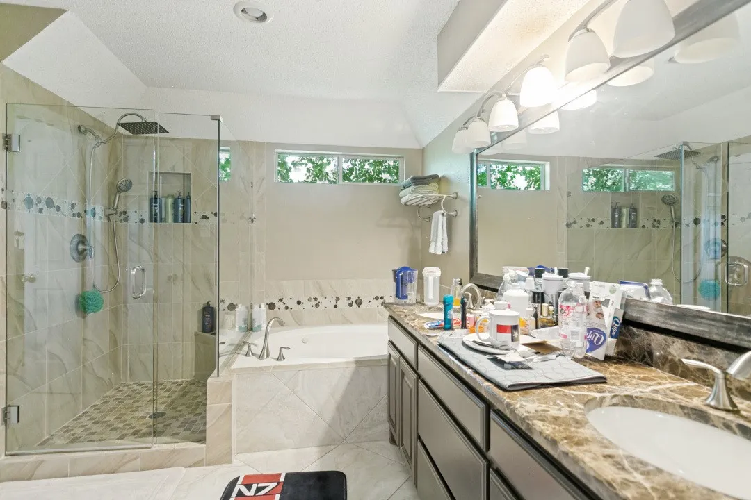Spacious bathroom featuring a glass-enclosed walk-in shower with a rain showerhead, a separate built-in soaking tub, a double vanity with an integrated mirror, warm-toned countertops, and multiple transom windows