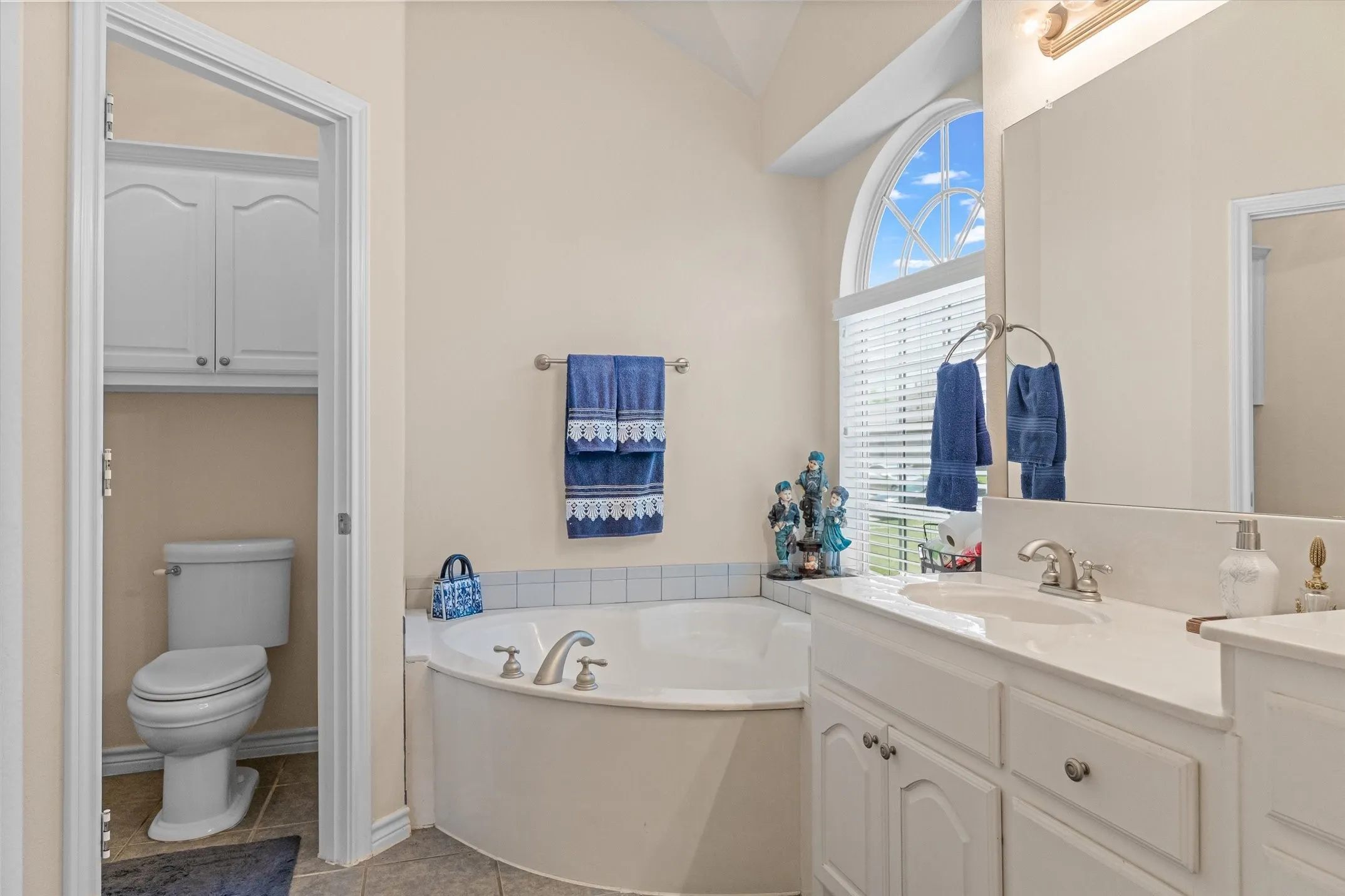 Soaking tub with tile surround, arched window, double vanity with built-in cabinetry, and a private water closet with additional built-in storage