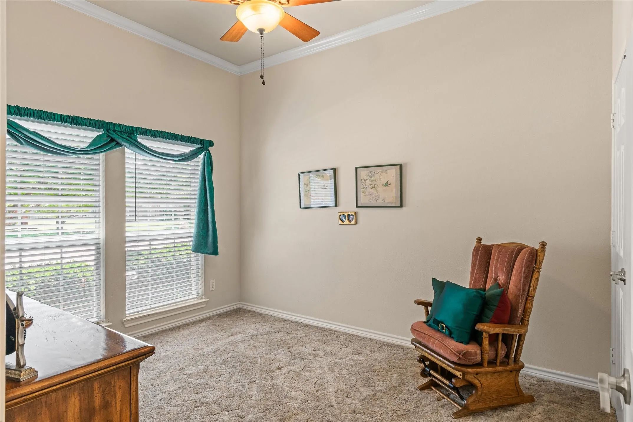 Vaulted ceiling with crown molding, ceiling fan with integrated lighting, double windows with blinds, light tan wall paint, and gray textured carpet