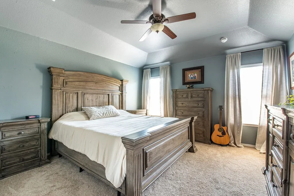 Carpeted bedroom with a tray ceiling, ceiling fan, and multiple windows with drapery