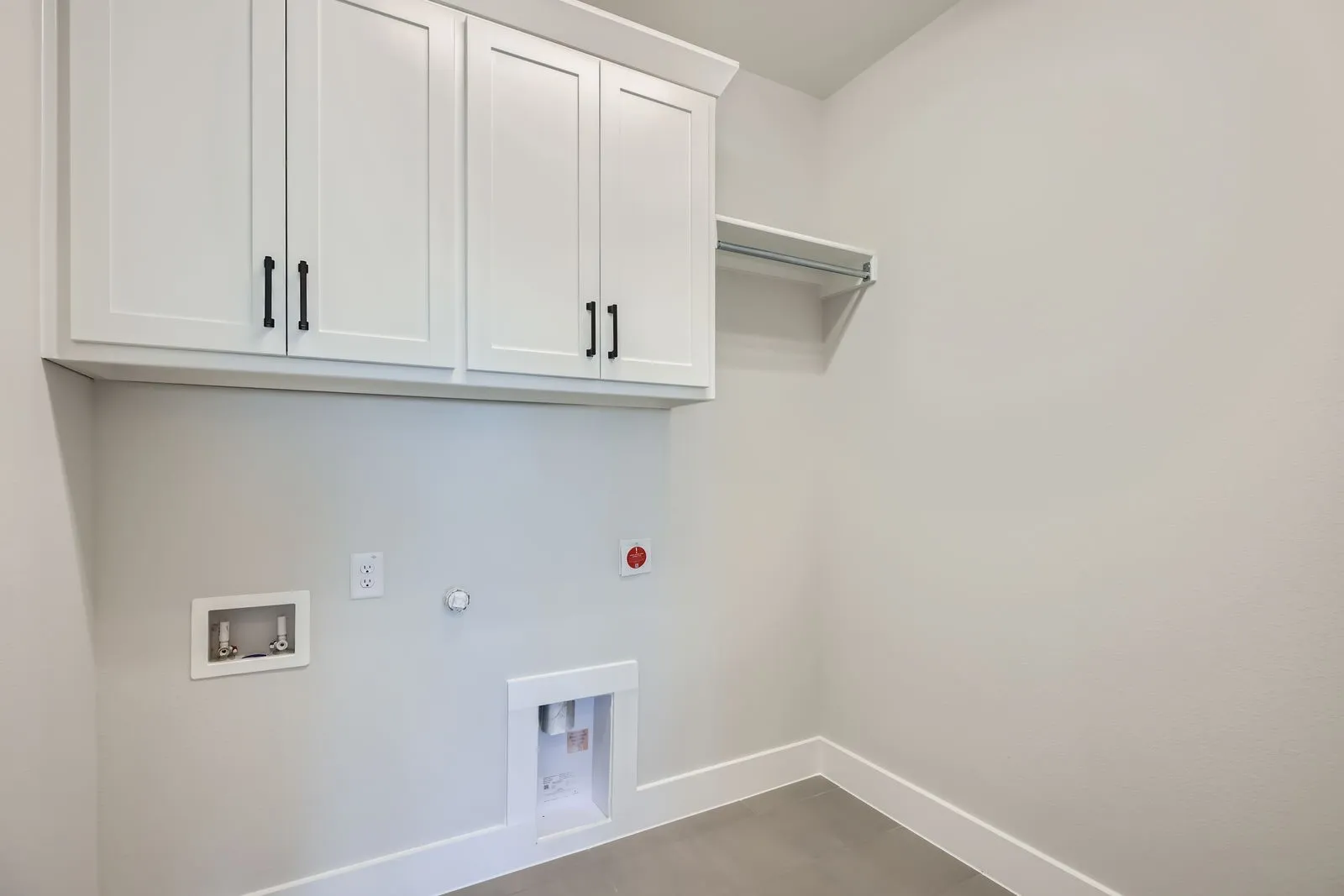 Laundry area with shaker-style upper cabinetry and matte black hardware, featuring a clothes rod, light gray flooring, and white baseboards