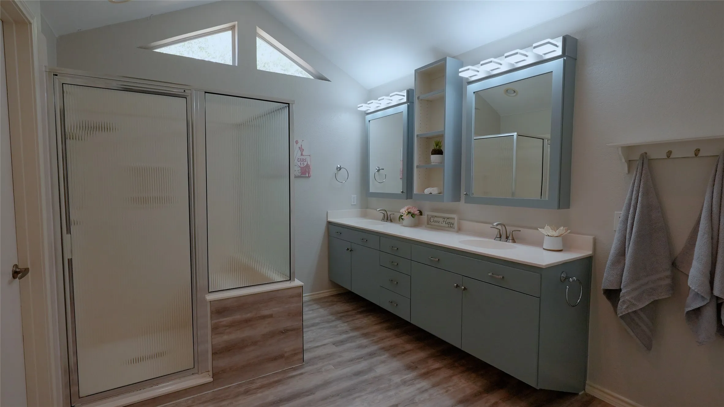 Vaulted ceiling with triangular windows, dual vanity with undermount sinks, wood-finish flooring, built-in cabinetry, and a glass-enclosed shower