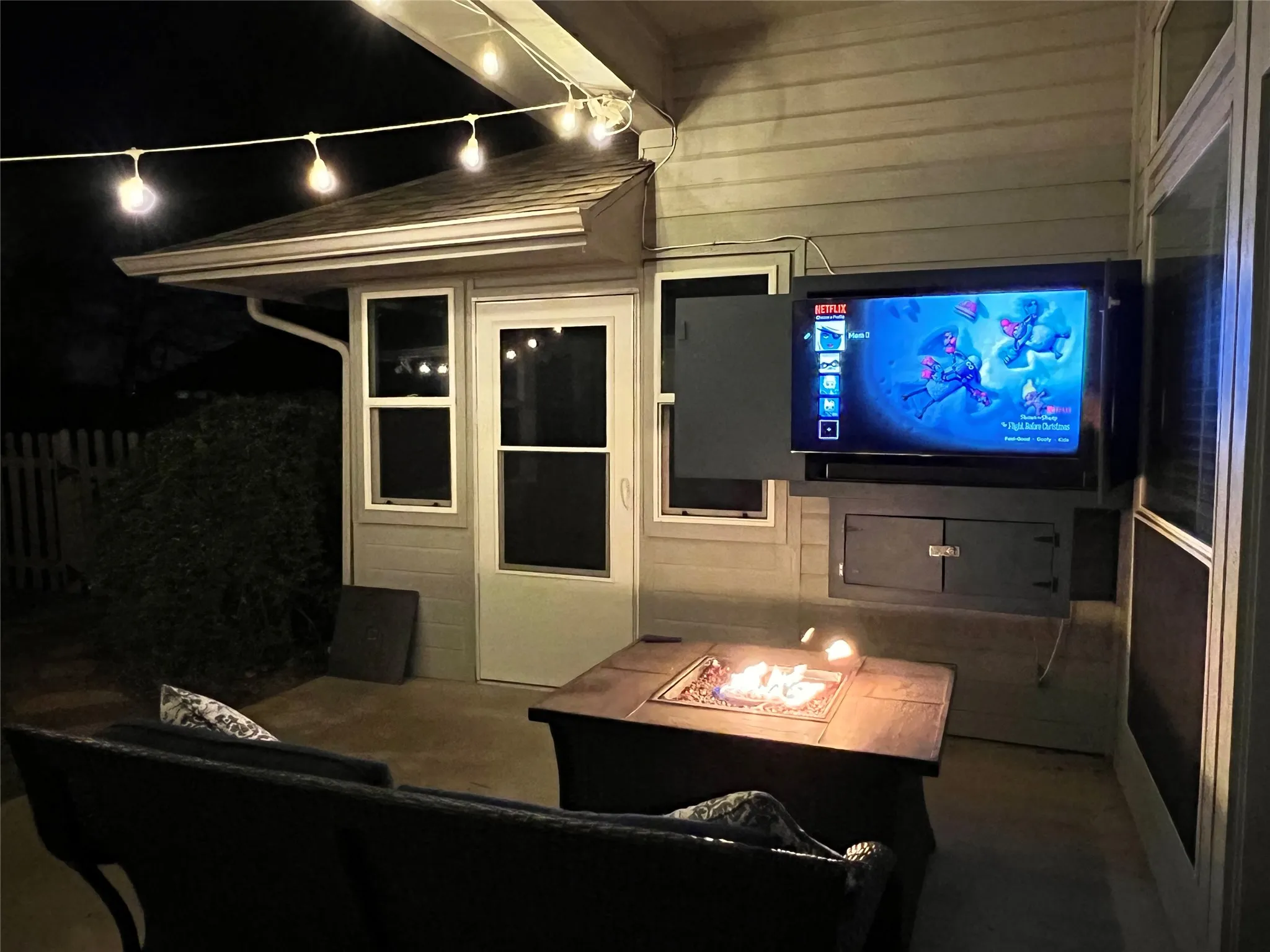 Covered patio featuring an exterior-mounted television, a built-in fire pit, and string lighting