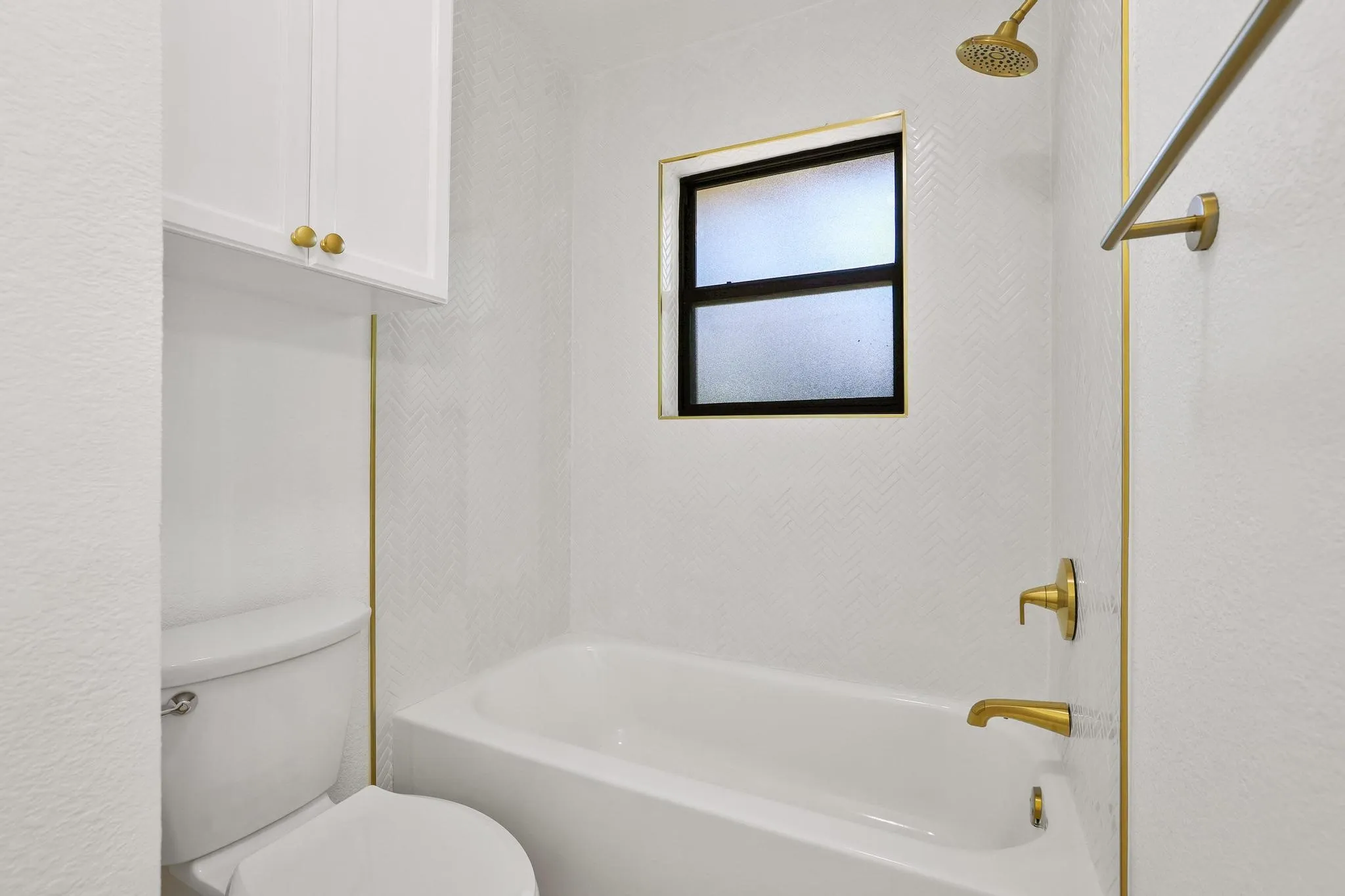 Shower-over-tub design with chevron-patterned tile, matte black window frame, and brushed gold fixtures