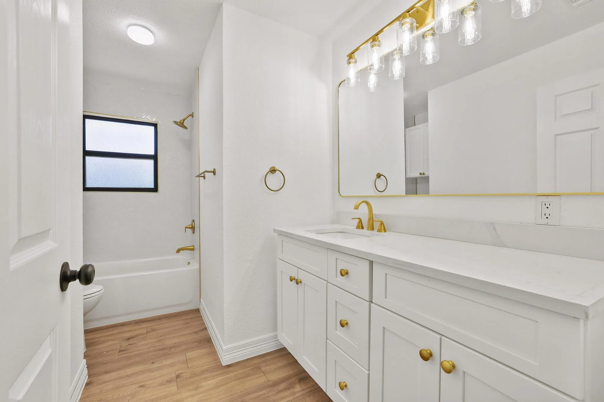 Bathroom featuring a wide vanity with white cabinetry and a light-toned countertop, a large mirror with a gold-toned frame, gold-toned fixtures, a bathtub with a shower, and wood-finish flooring