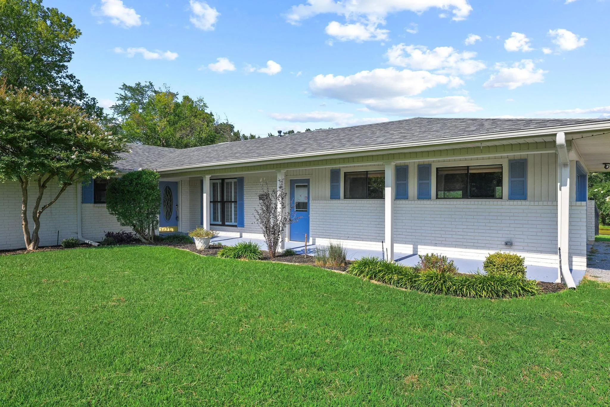 Ranch-style residence featuring white brick and siding, a covered entry, blue accent shutters, and a well-maintained lawn with mature landscaping