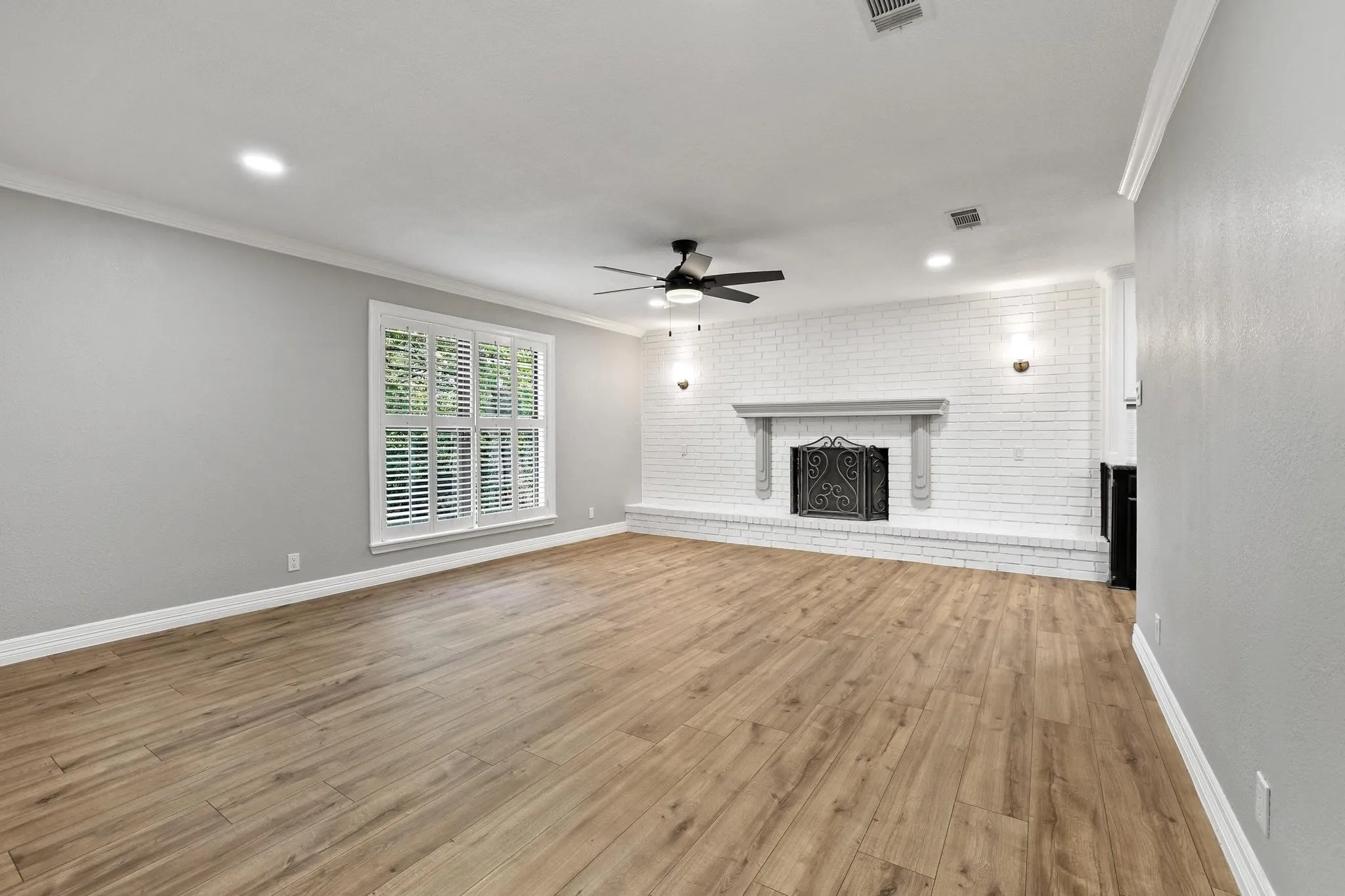 Spacious room featuring wood-finish flooring, a white-painted brick fireplace with a detailed screen, and a large window with white plantation shutters