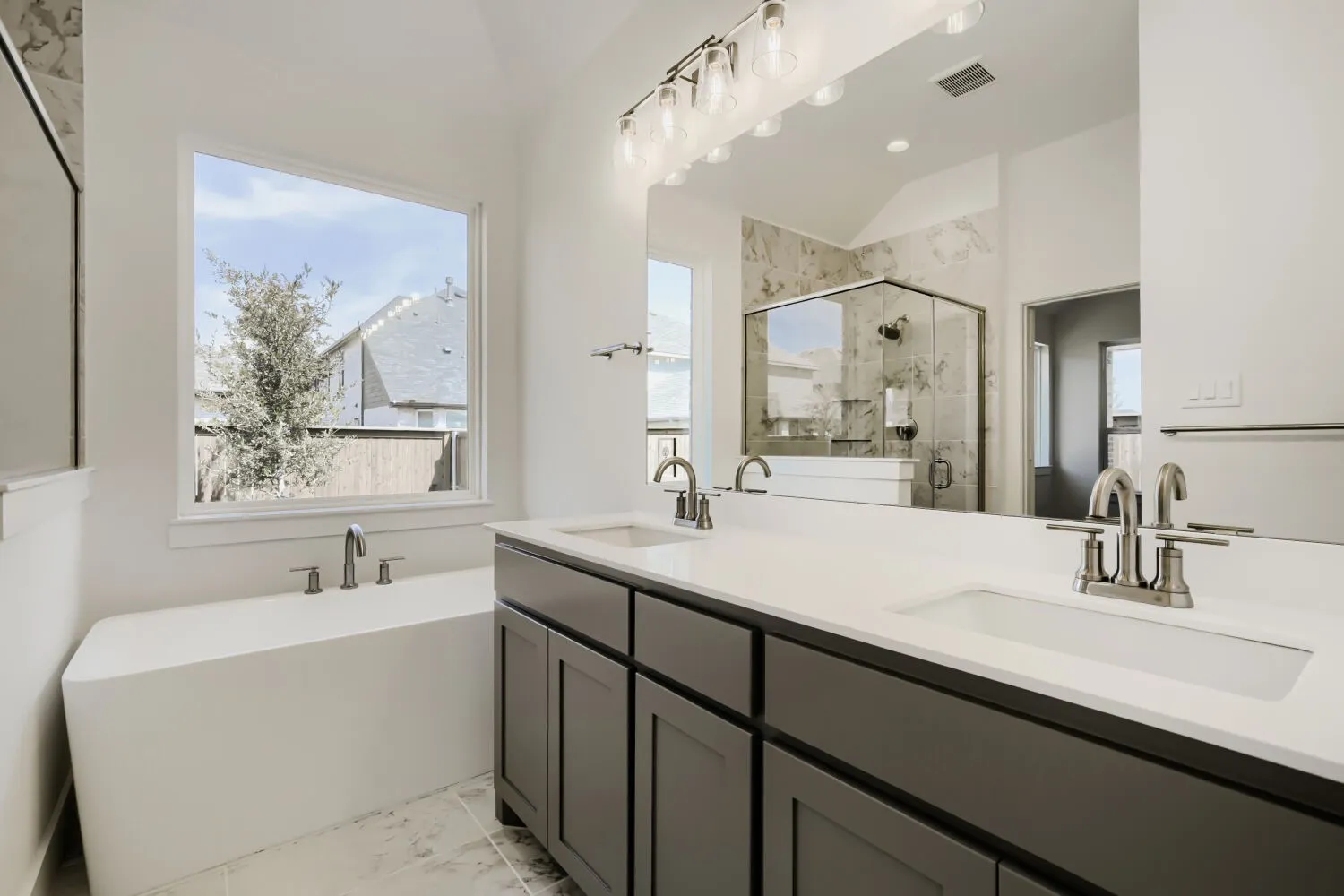 Dual vanity with integrated sinks and brushed nickel fixtures, a frameless glass shower enclosure, a freestanding soaking tub, and a large window