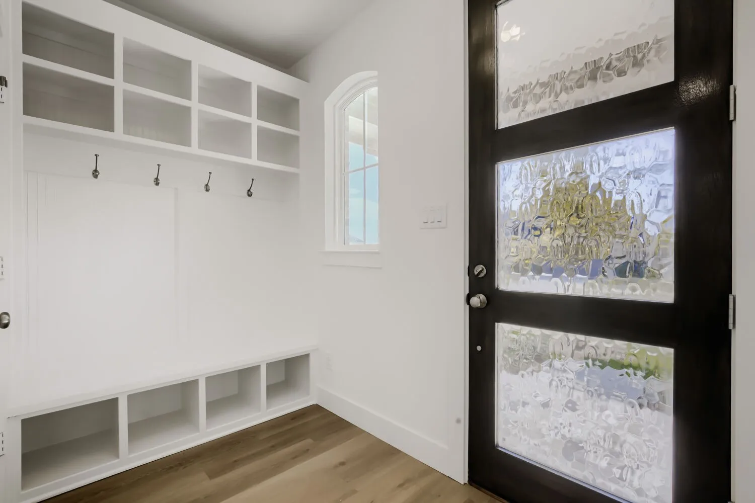 Dark-finish entry door with textured glass panels, built-in storage bench with cubbies, upper storage cubbies, decorative wall hooks, and wood-finish flooring