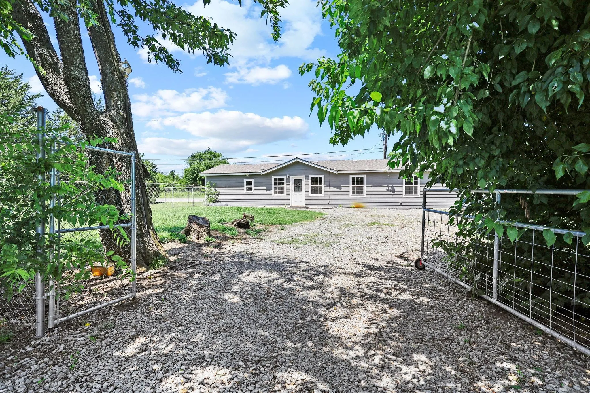 Gravel driveway with two metal gates opening to a single-story residence featuring grey siding, white trim, and multiple windows