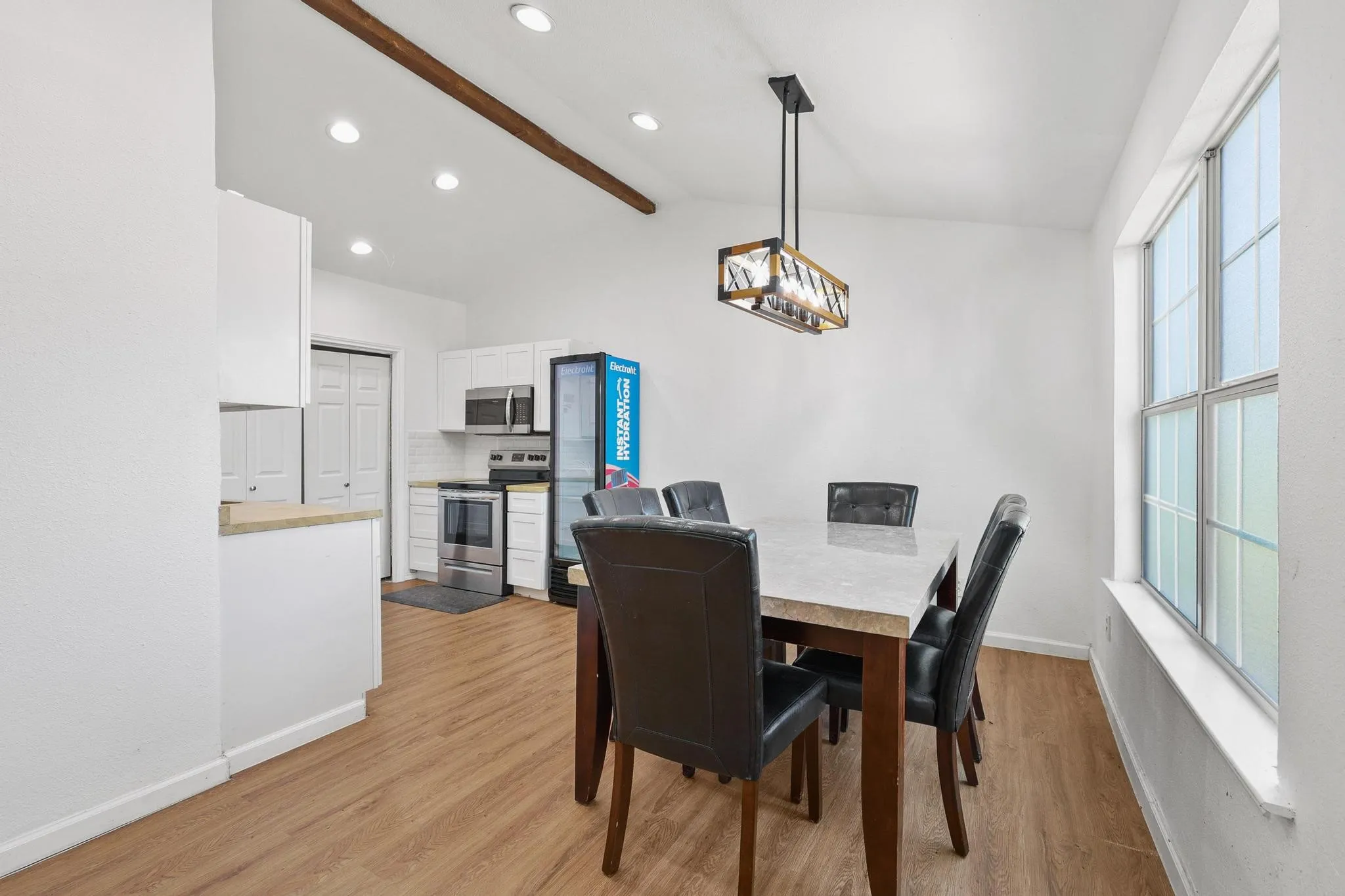 Open-concept kitchen and dining area featuring vaulted ceilings with exposed wood beams, wood-finish flooring, a contemporary linear chandelier, and a large multi-pane window