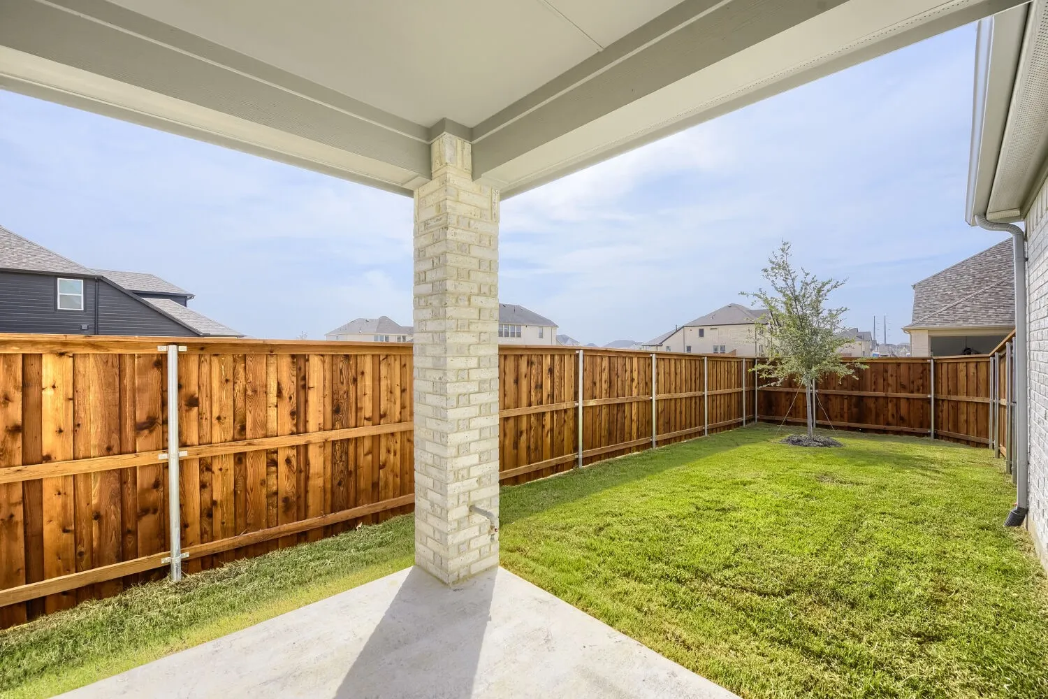 Covered patio with a brick support column, concrete flooring, and a fully fenced backyard with a single tree