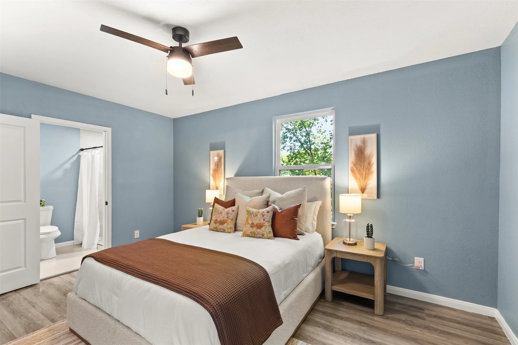 Bedroom featuring wood-finish flooring, a ceiling fan with integrated lighting, a window with exterior views, and an en-suite bathroom with a toilet and shower