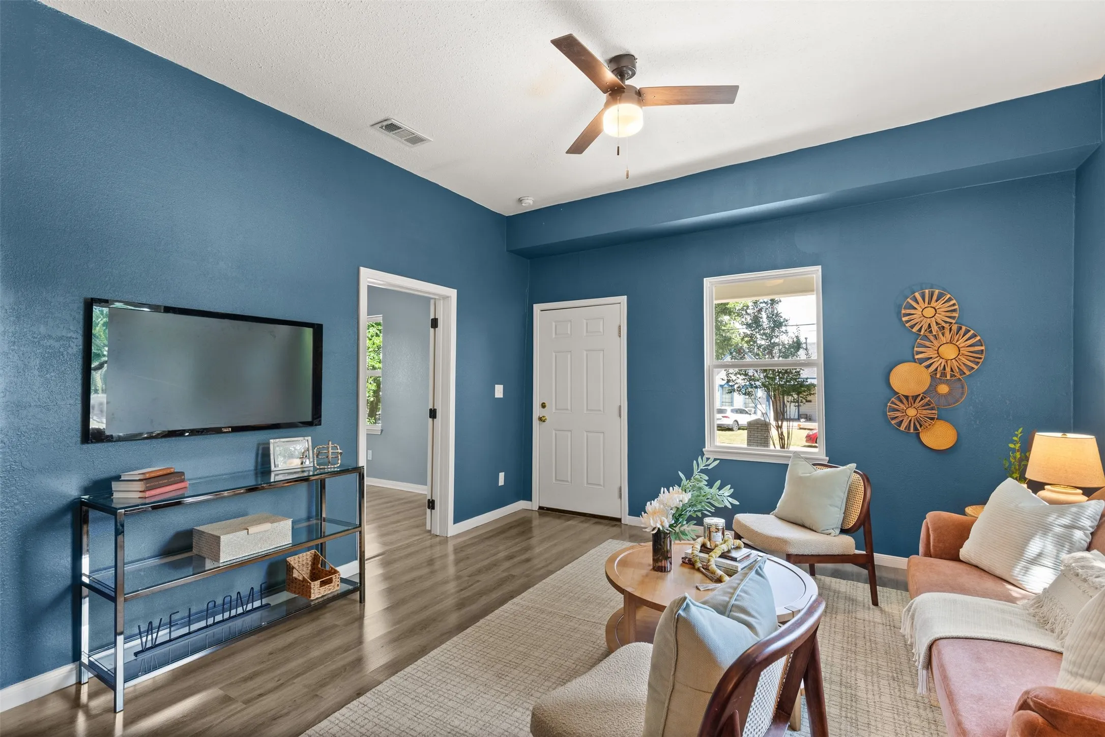 Spacious living area featuring plank wood-finish flooring, a ceiling fan with integrated lighting, and a white paneled entry door