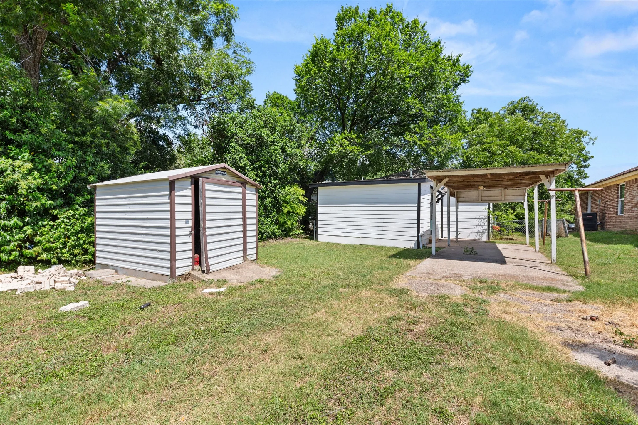 Expansive backyard featuring a detached storage shed, a covered carport with a concrete pad, and mature trees providing shade