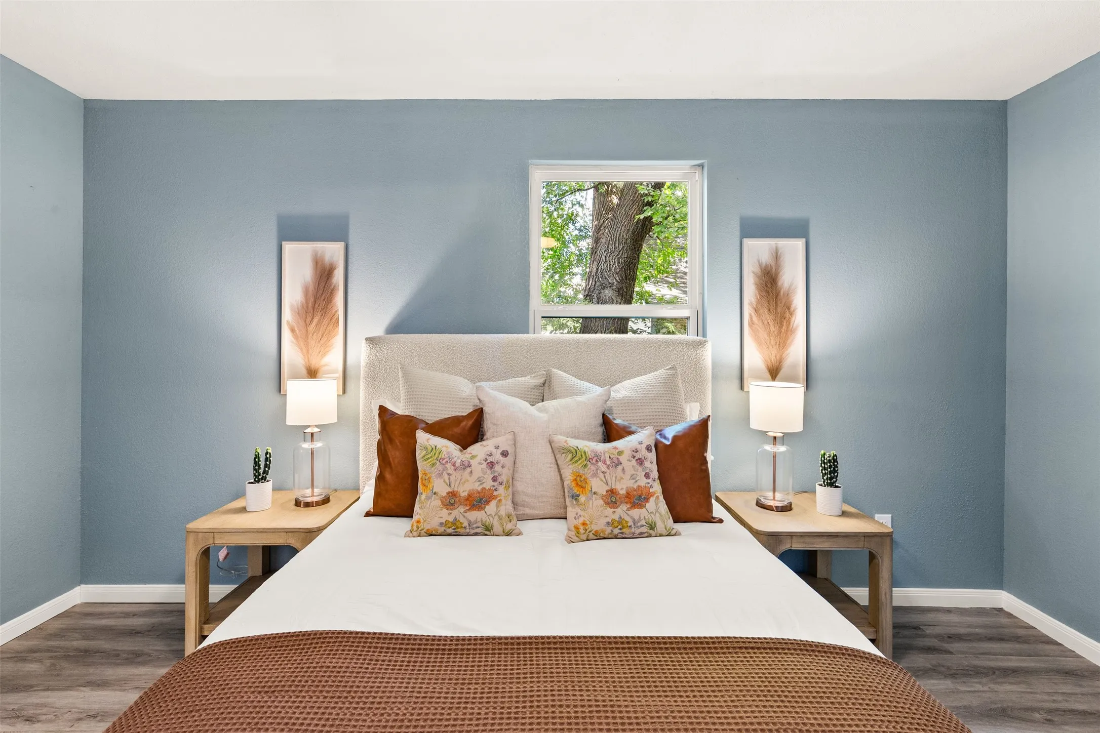 Bedroom featuring light blue walls, wood-finish flooring, and a central window overlooking mature trees