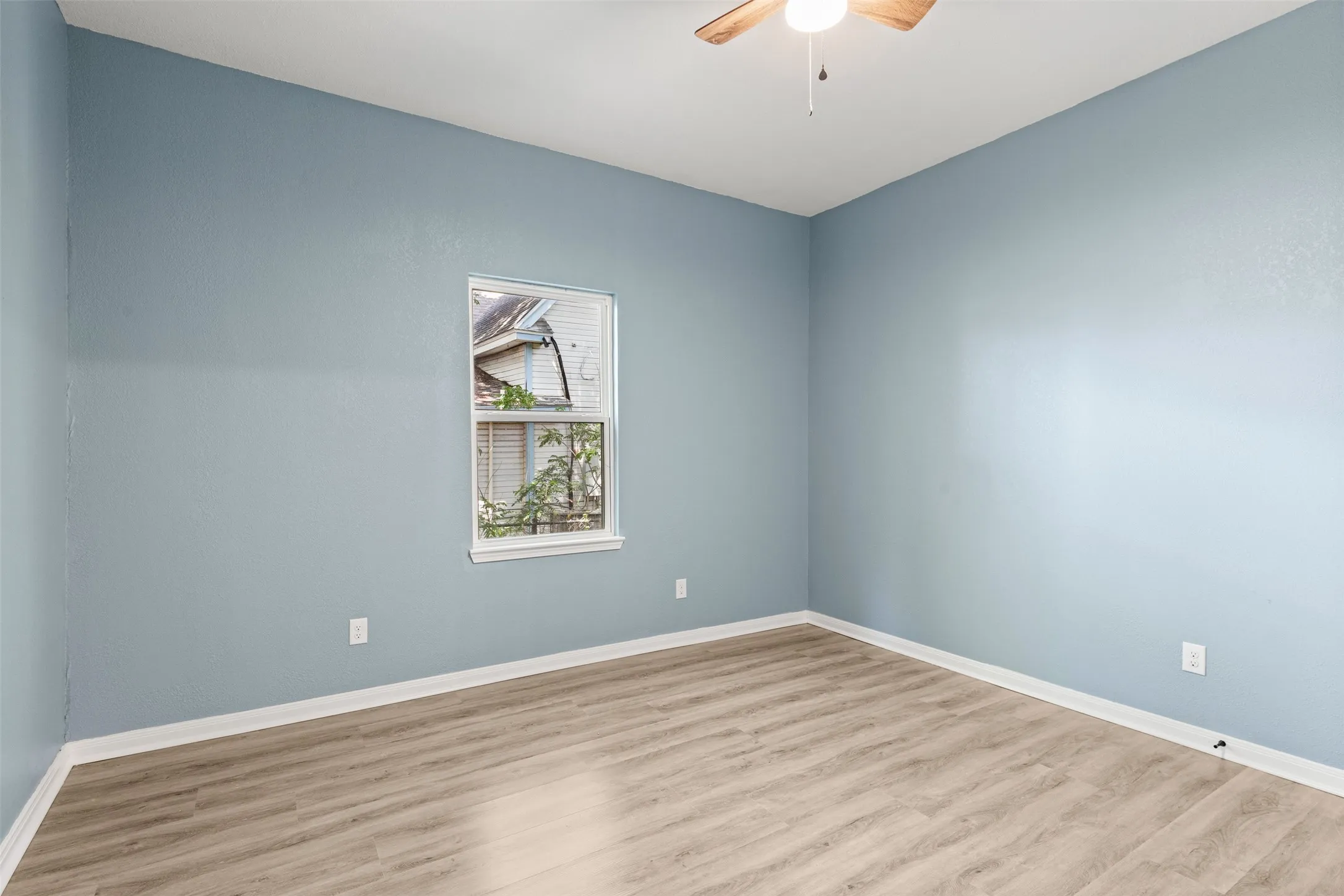 Room featuring light blue walls, wood-finish flooring, a white-framed window, white baseboards, and a ceiling fan with integrated lighting