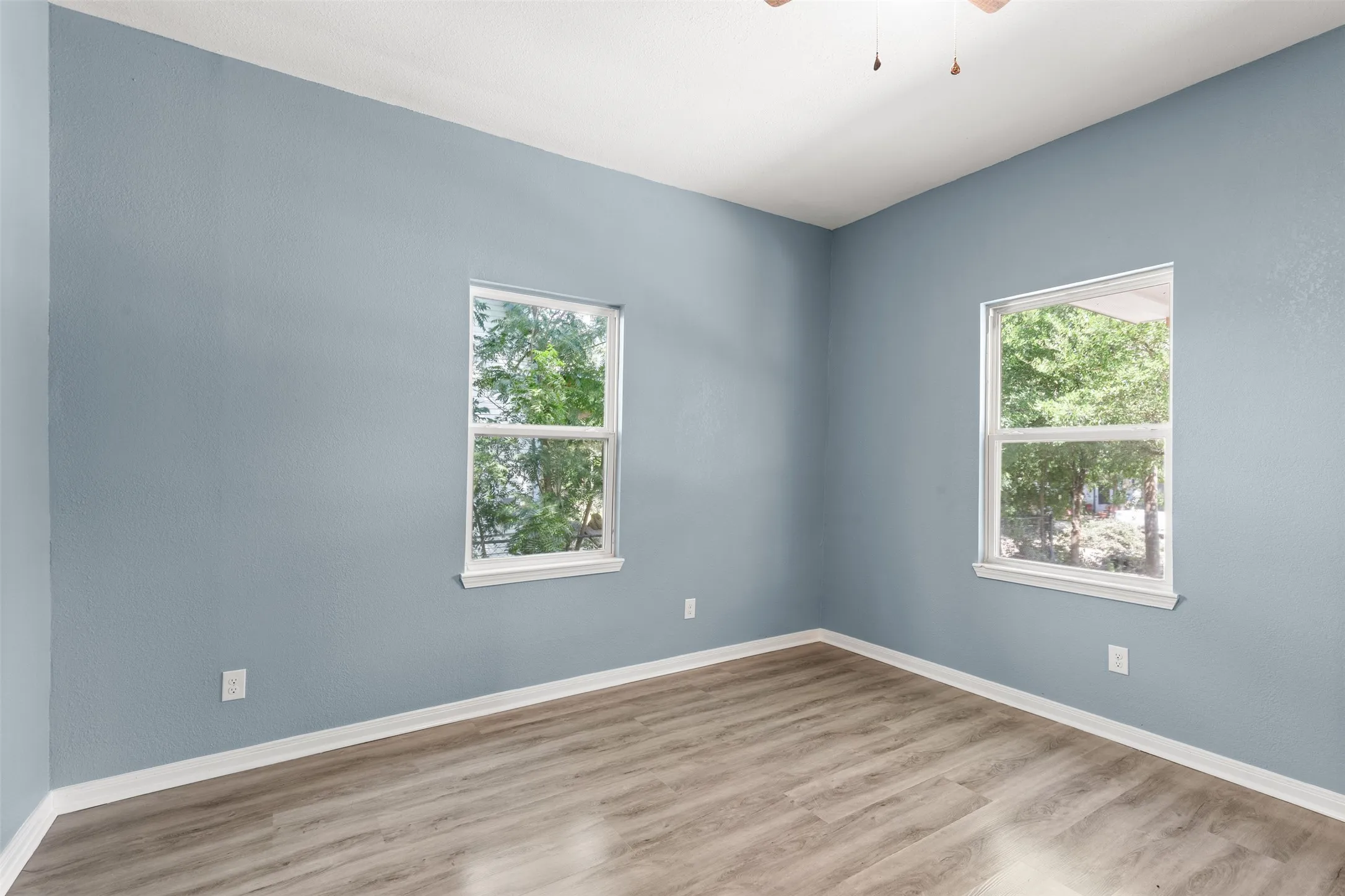 Room featuring wood-finish flooring, light blue walls, two windows with white trim, and a ceiling fan