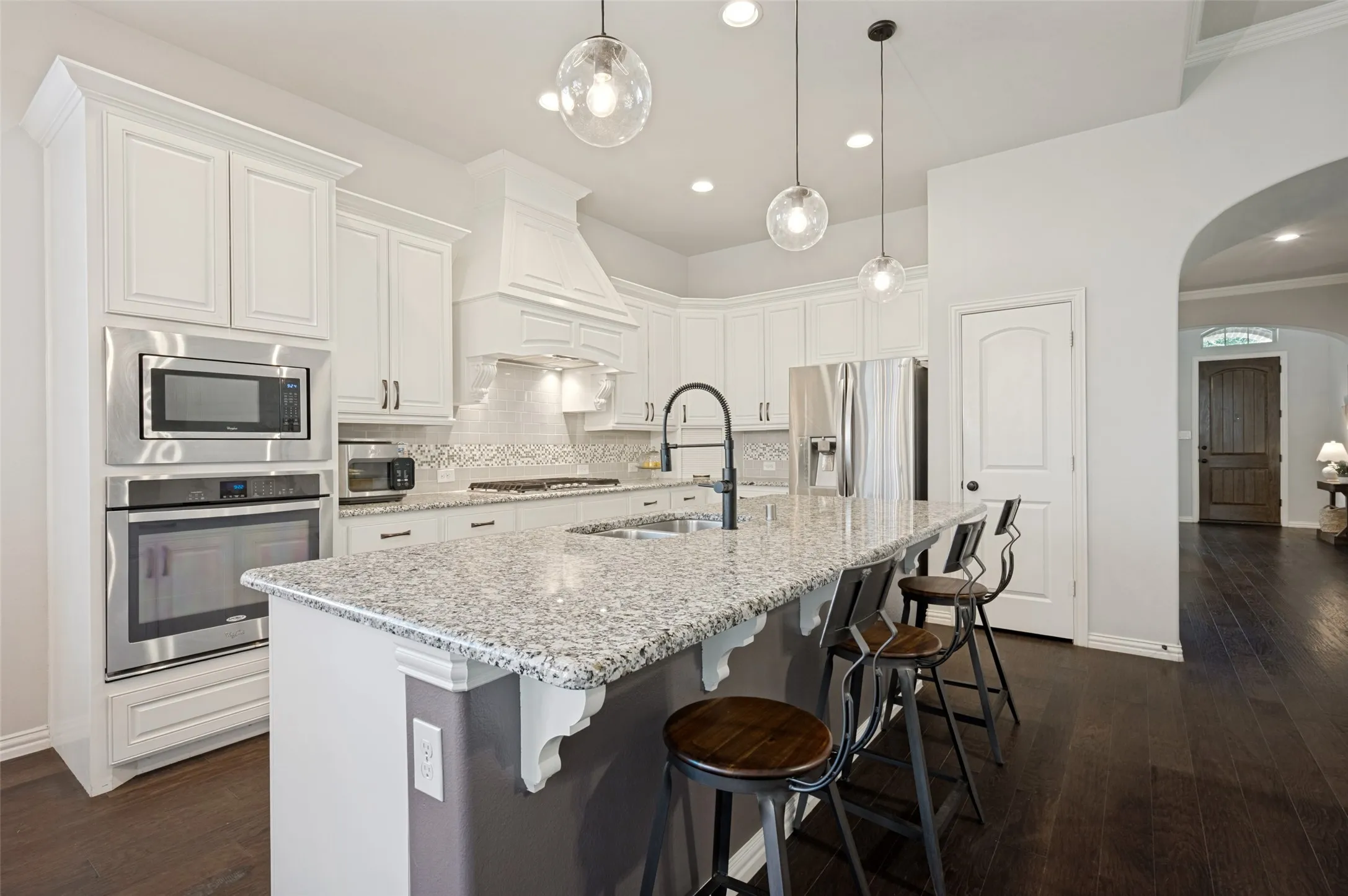 Spacious kitchen featuring an expansive island with a granite countertop, white cabinetry, integrated stainless steel appliances, a tile backsplash, and dark wood-finish flooring