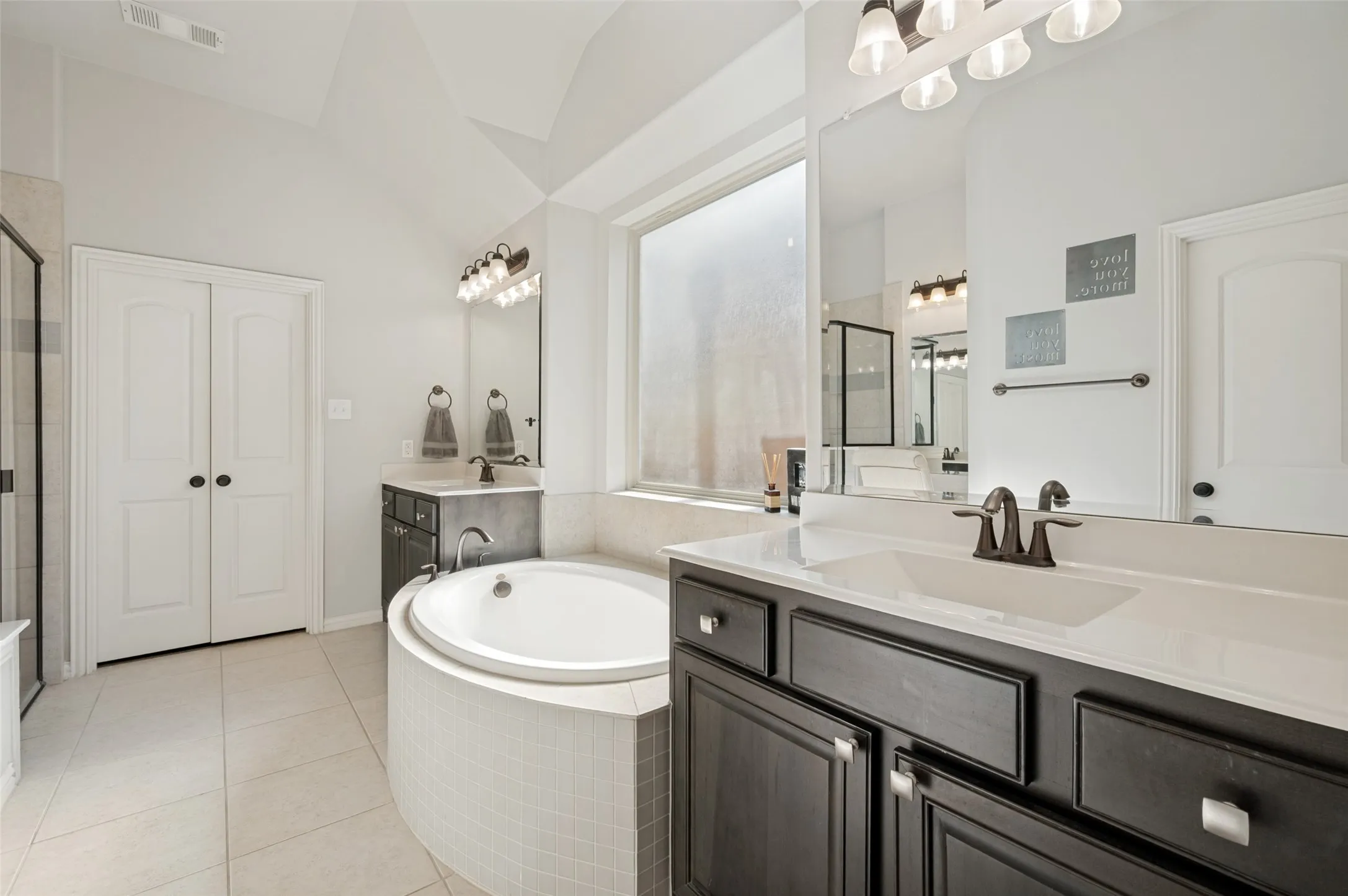 Vaulted ceiling bathroom featuring a built-in soaking tub with mosaic tile surround, dual dark wood-finish vanities with integrated sinks, and a large privacy window