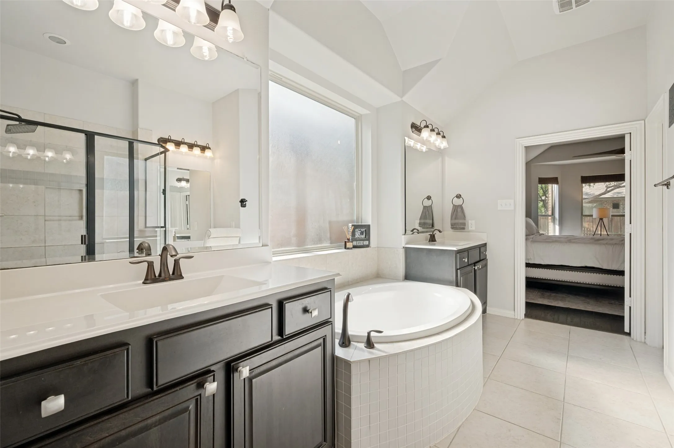 Bathroom featuring a large soaking tub with tile surround, dual vanities with white countertops and dark cabinetry, a separate glass-enclosed shower, and vaulted ceilings