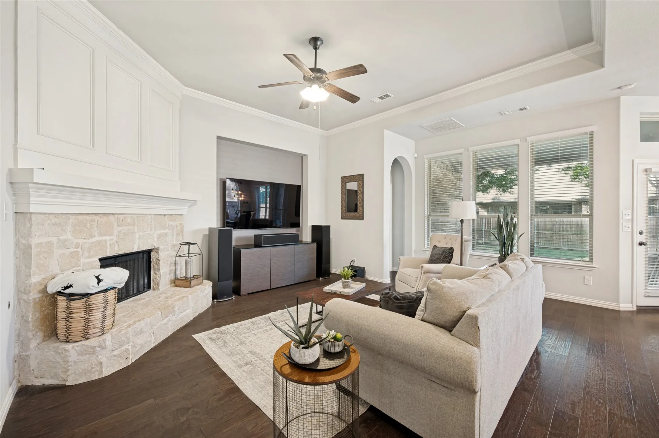 Spacious living area featuring dark wood-finish flooring, a stone-surround fireplace with a white mantel, multiple large windows, a tray ceiling, and a ceiling fan