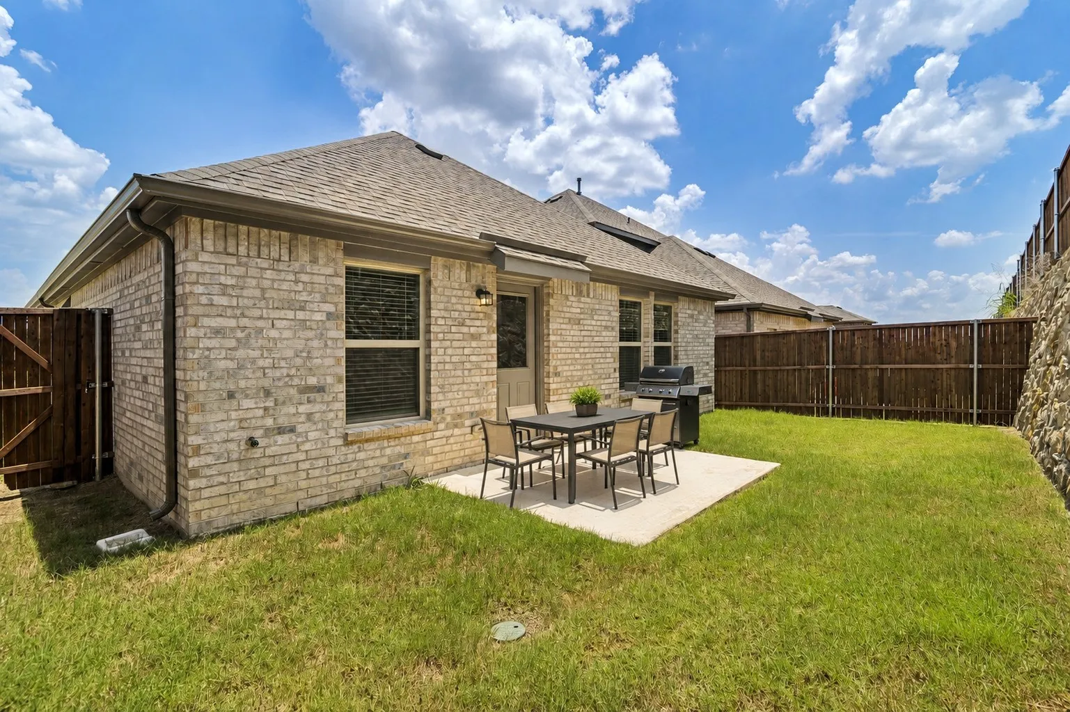Brick exterior with covered patio featuring concrete flooring and metal dining furniture, backing to a spacious lawn with wooden privacy fencing