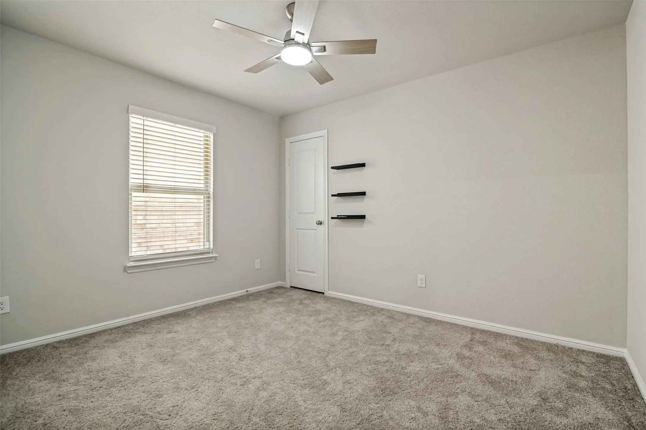 Room featuring light gray walls, gray carpet flooring, a window with blinds, a ceiling fan with integrated lighting, and a white door with black floating shelves