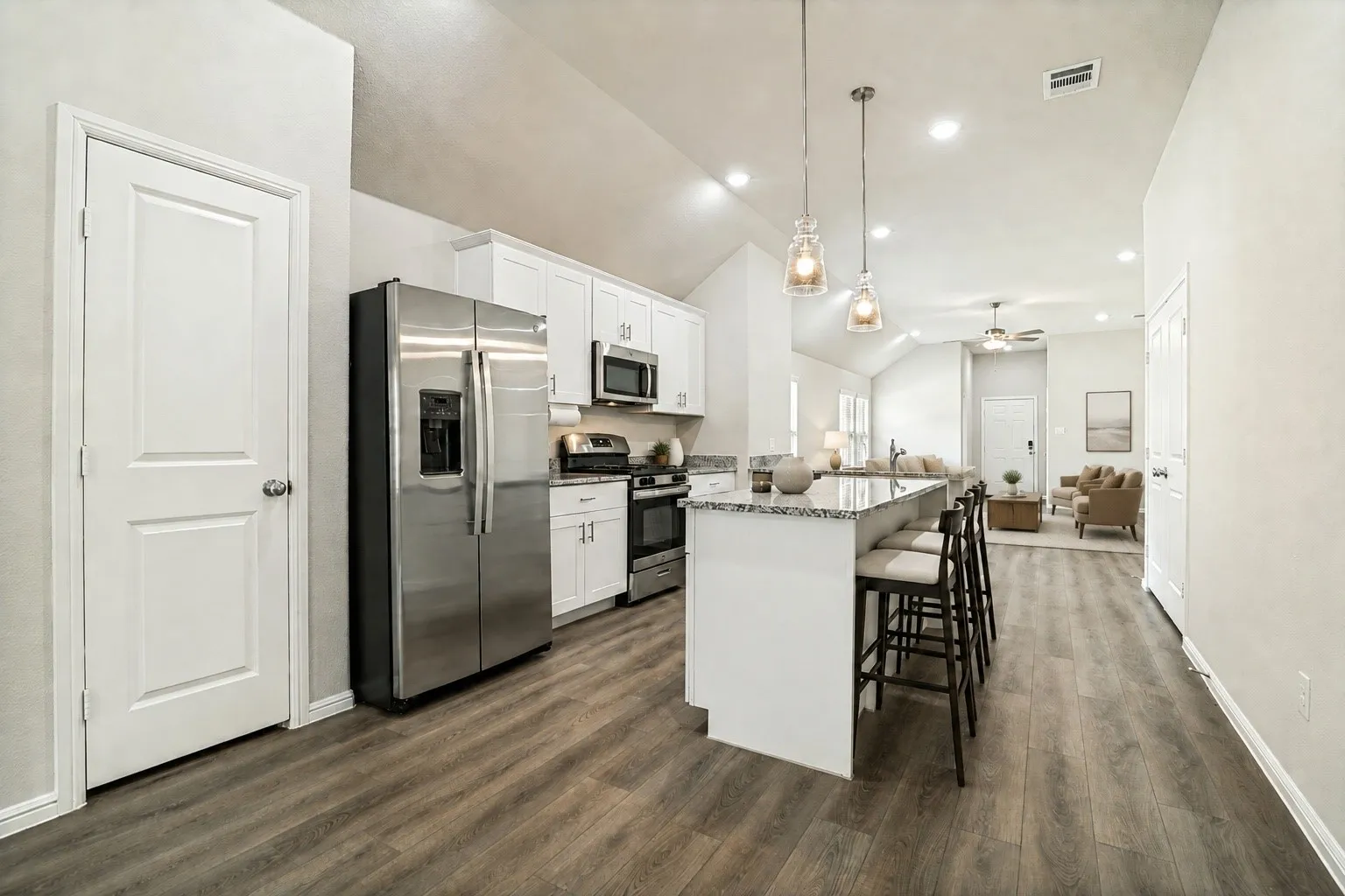 Modern open-concept kitchen and living area featuring stainless steel appliances, white cabinetry with hardware accents, a large waterfall-edge island with seating for multiple, wood-finish flooring throughout, and recessed ceiling lighting with pendant fixtures (virtually staged)