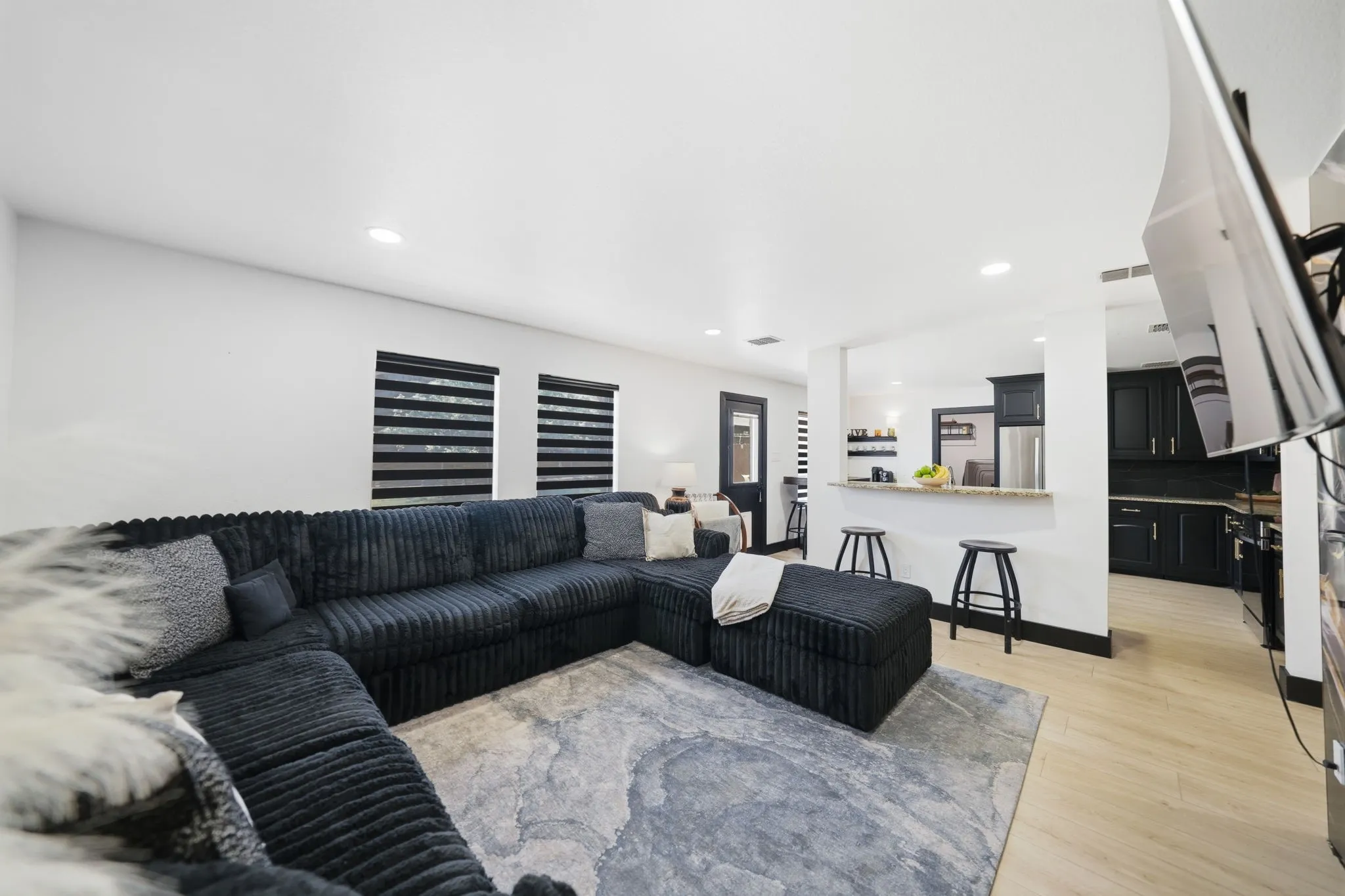 Open-concept living space featuring wood-finish flooring, recessed lighting, a breakfast bar with stone countertop, and a kitchen with black cabinetry and stainless steel appliances